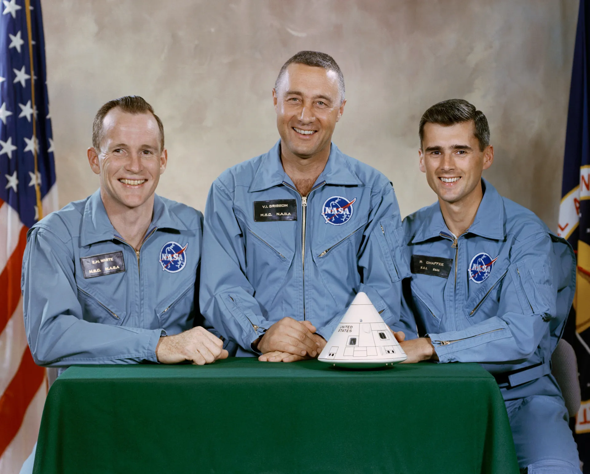 Ed White, Gus Grissom and Roger Chaffee in blue flight suits, seated at a table with a model of their command module.