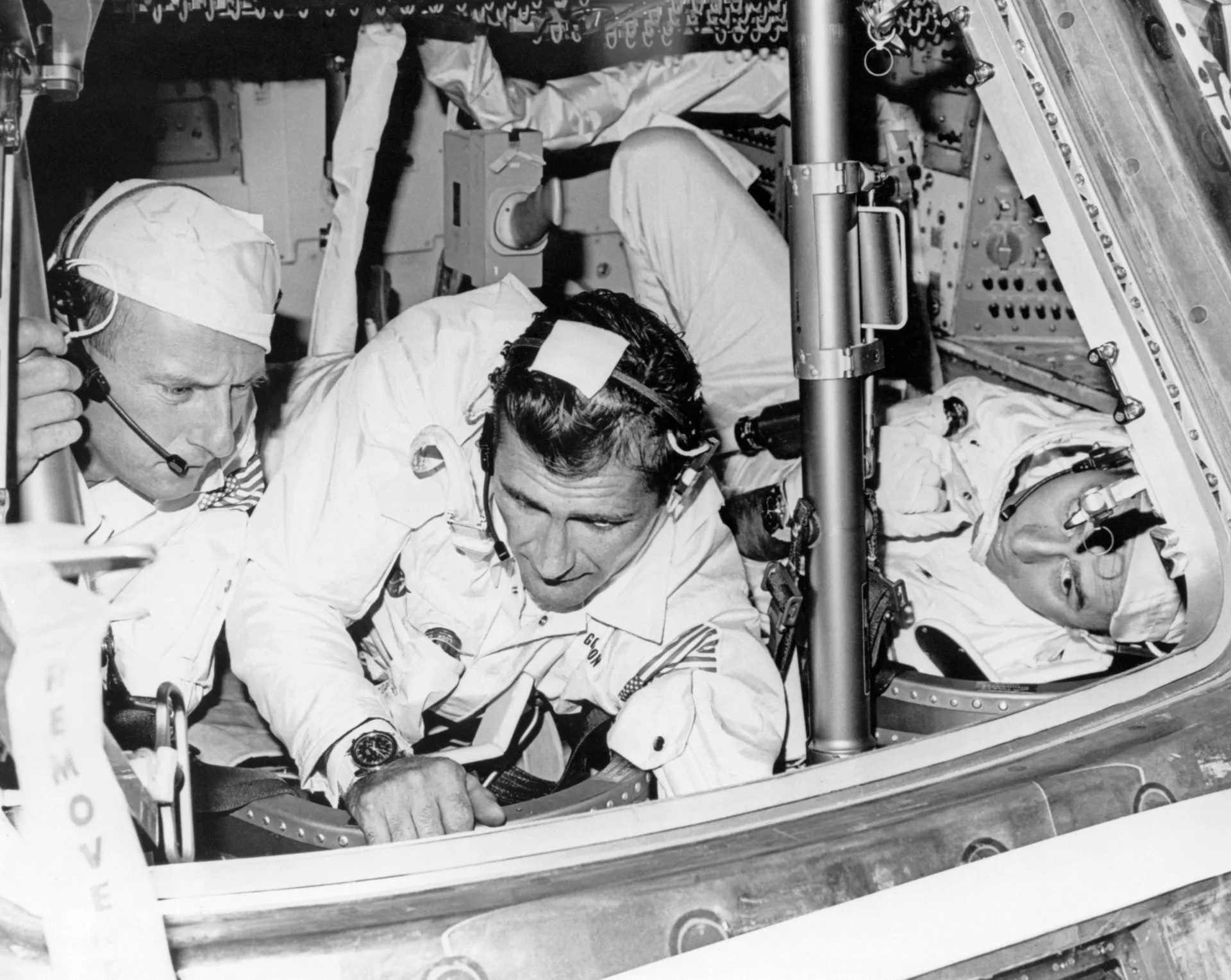 Three astronauts in white coveralls check out a command module, one lying in a couch, at the North American plant.