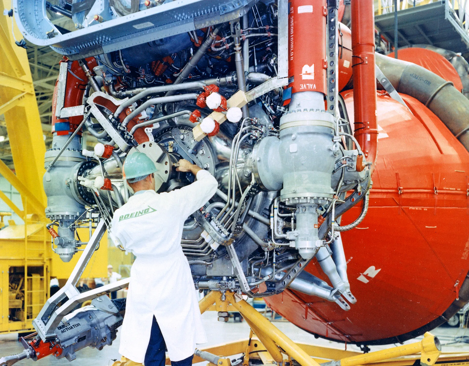 A Boeing technician in a white coat and green hard hat works among the pipes of an F-1 engine on a Saturn V first stage.