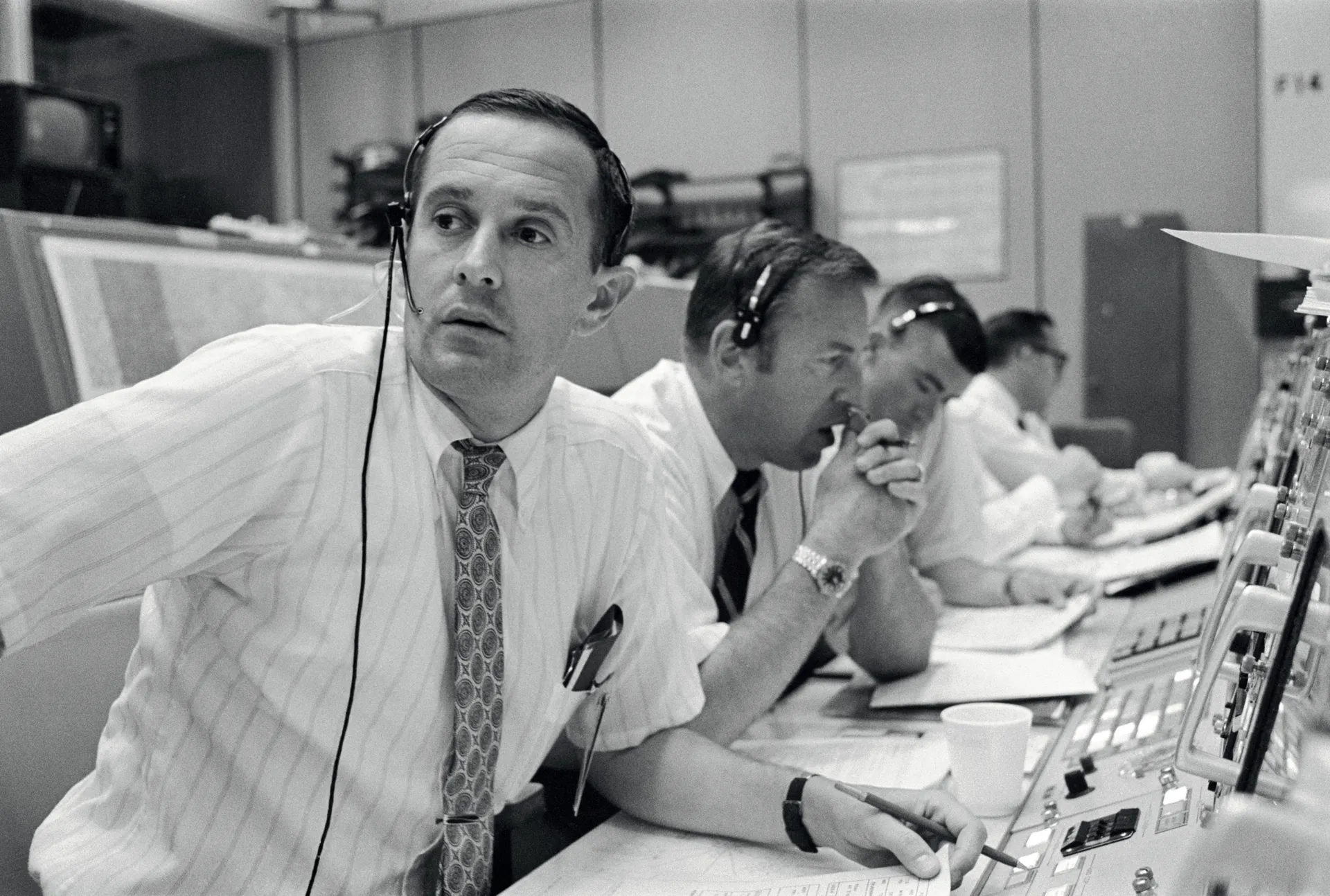Capcom Charles Duke, wearing a headset, watches his console intently, with Jim Lovell and Fred Haise beside him.