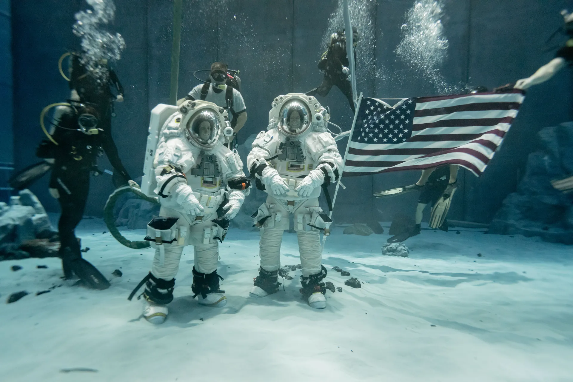 Two astronauts in white Axiom lunar spacesuits stand on the floor of NASA’s Neutral Buoyancy Laboratory pool beside a U.S. flag, with divers around them.