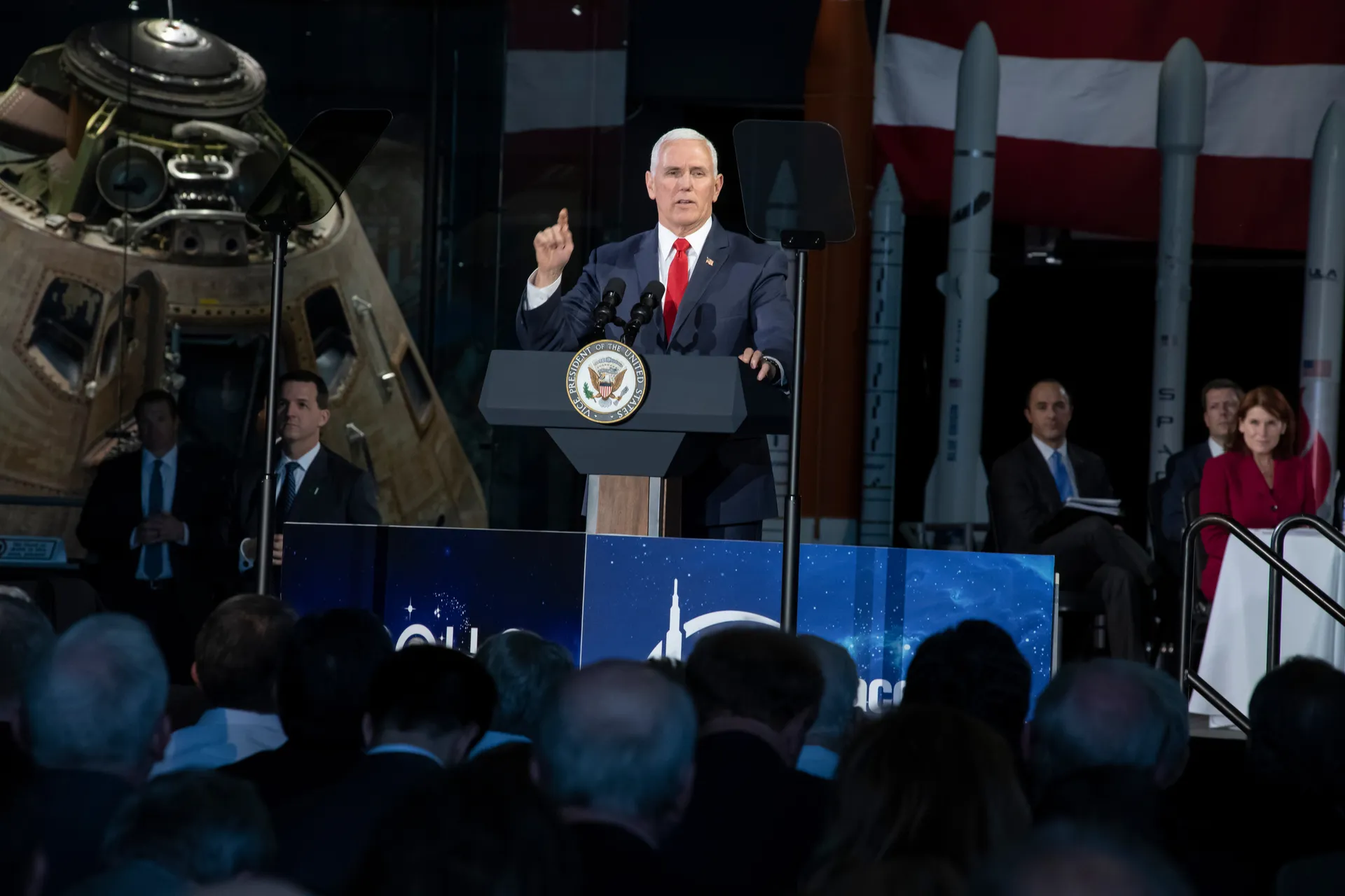 Vice President Mike Pence speaks at a lectern with an Apollo command module behind him on the left and rocket models on the right; an audience fills the foreground.