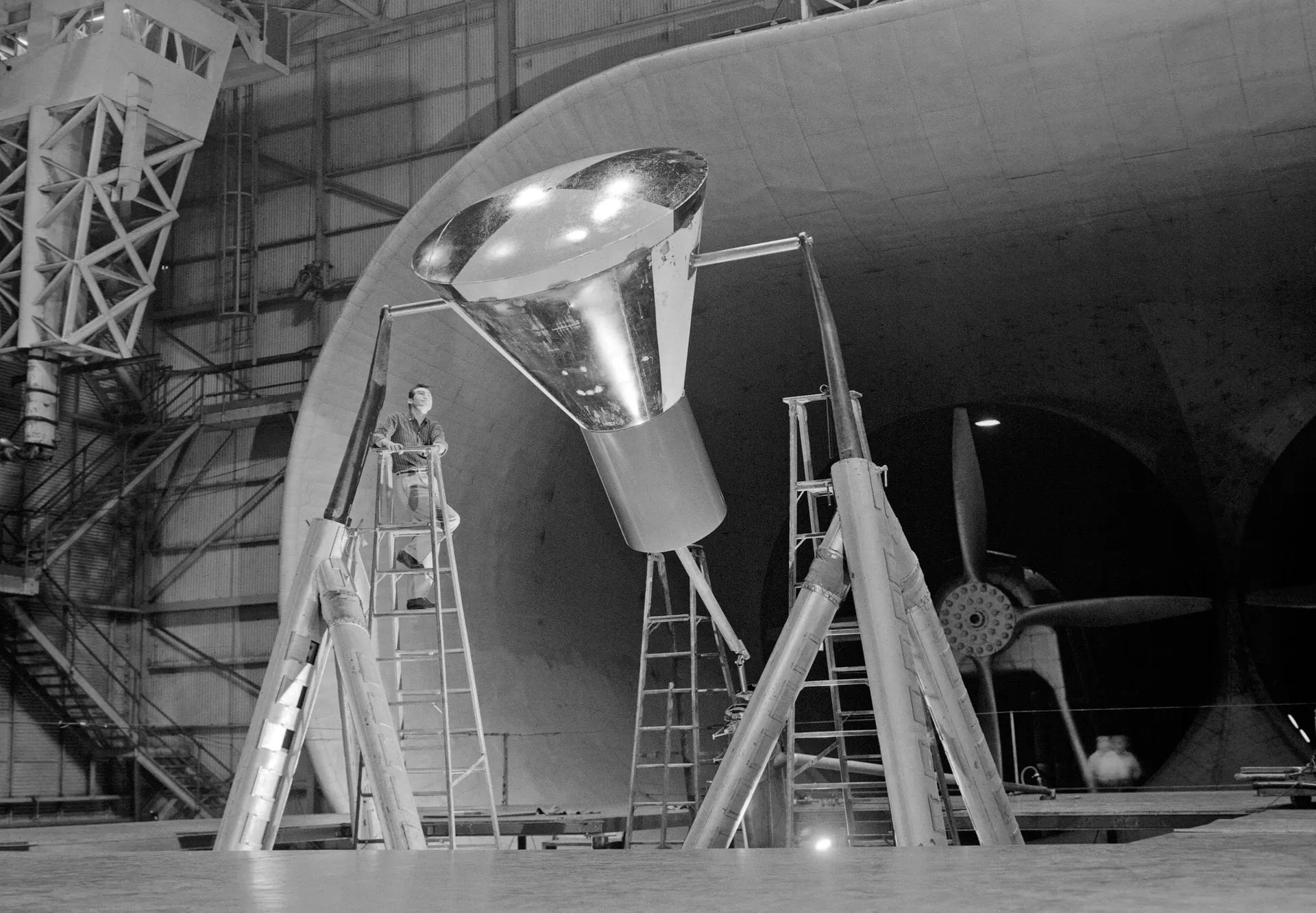 A full-size Mercury capsule mounted on struts inside Langley’s Full Scale Tunnel, with a technician on a ladder and the tunnel’s giant fan behind.
