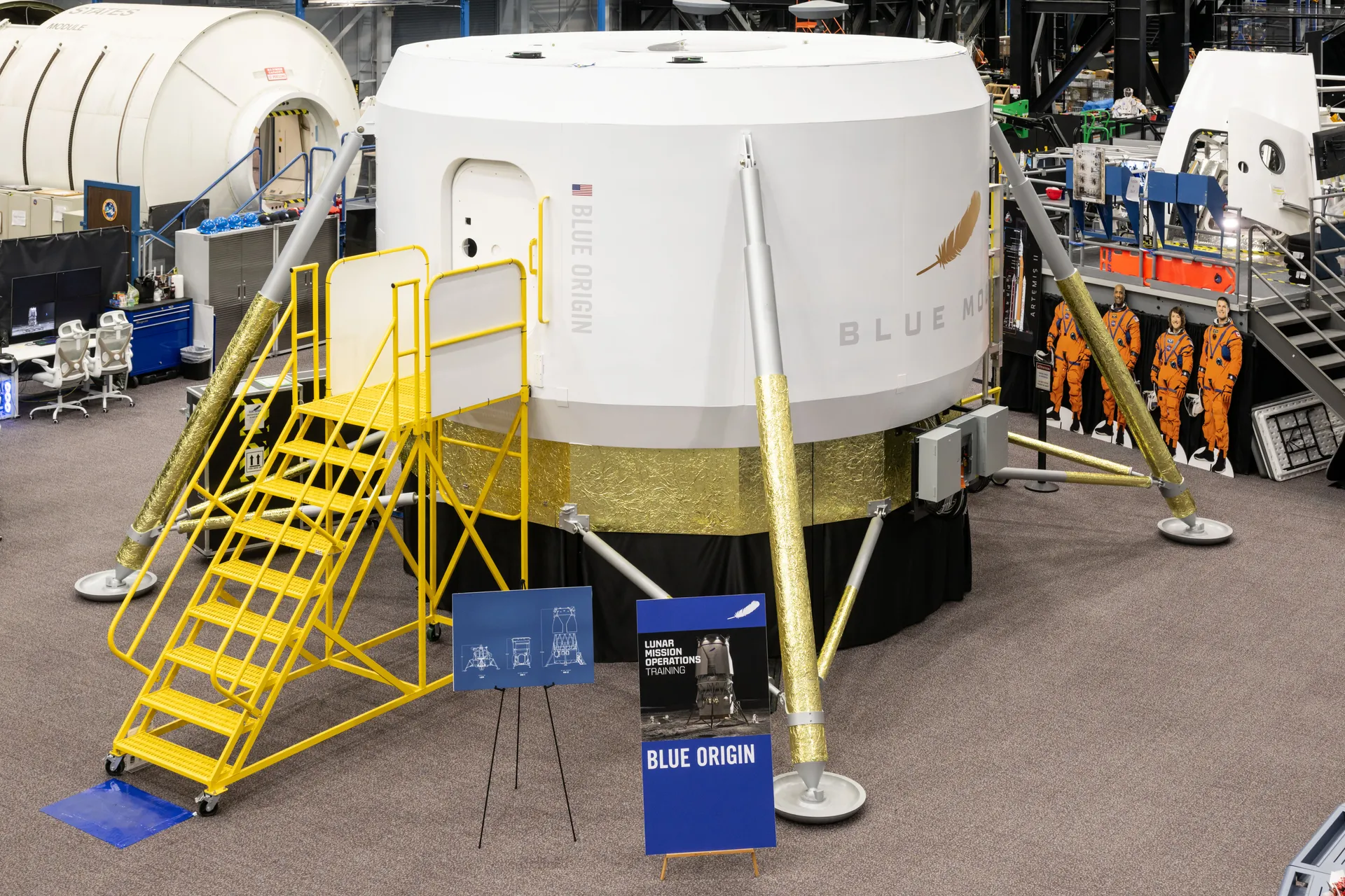 A full-size white crew cabin mockup on four gold-wrapped legs, with a yellow work stand leading to its hatch, in a training hall.