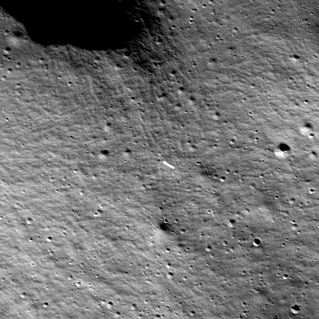 An overhead view of gray, cratered lunar terrain with a small white arrow pointing to the lander.
