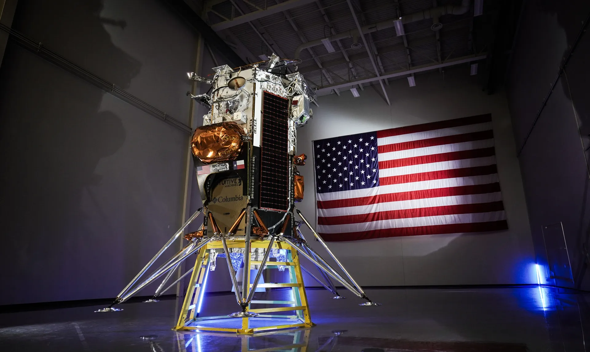 A six-legged, hexagonal lunar lander with gold and silver panels stands in a dark hangar before an American flag.