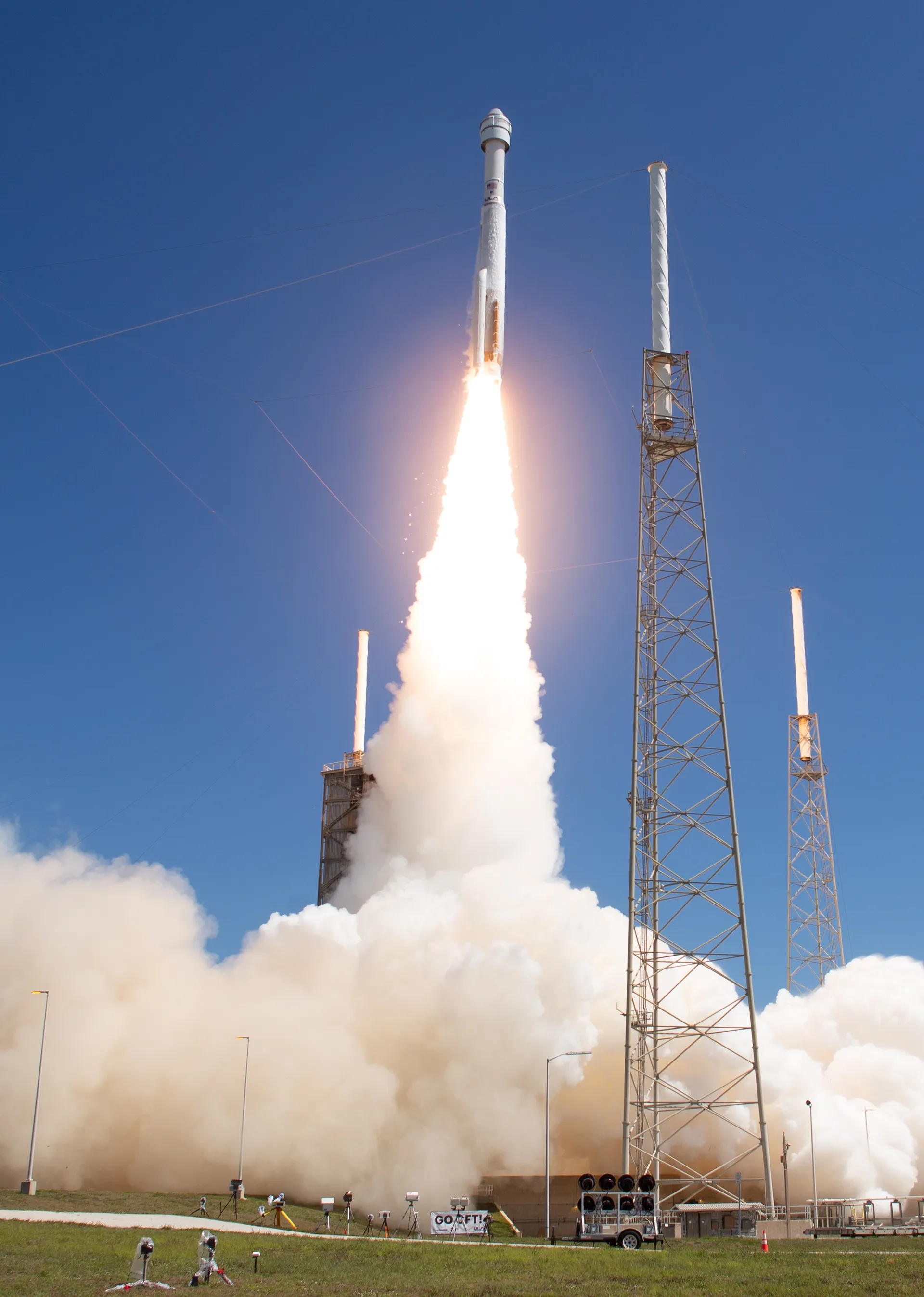 An Atlas V rocket climbs on a bright plume past lightning towers against a blue sky.