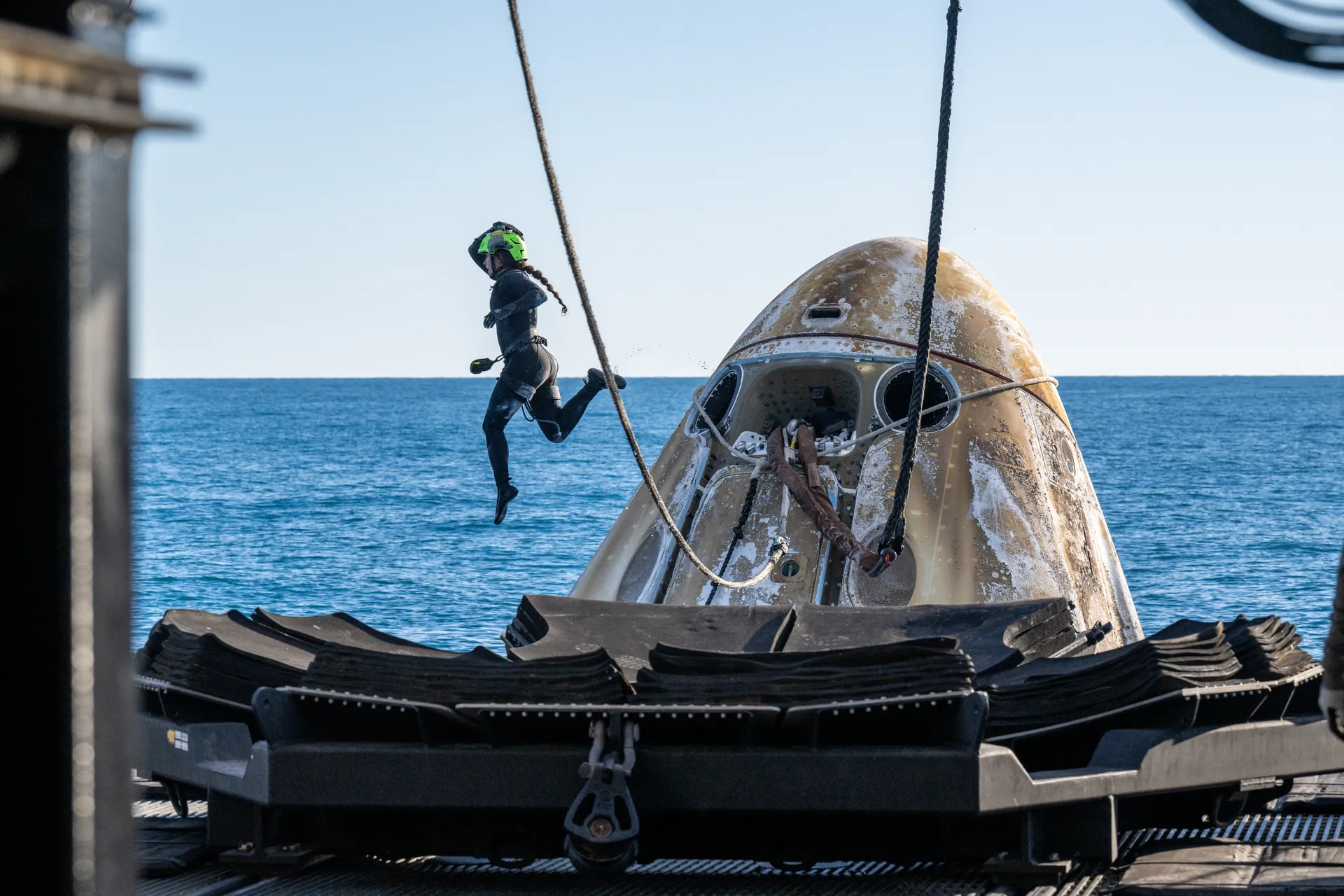 A recovery swimmer leaps from a ship’s deck toward a scorched white capsule floating in blue water.