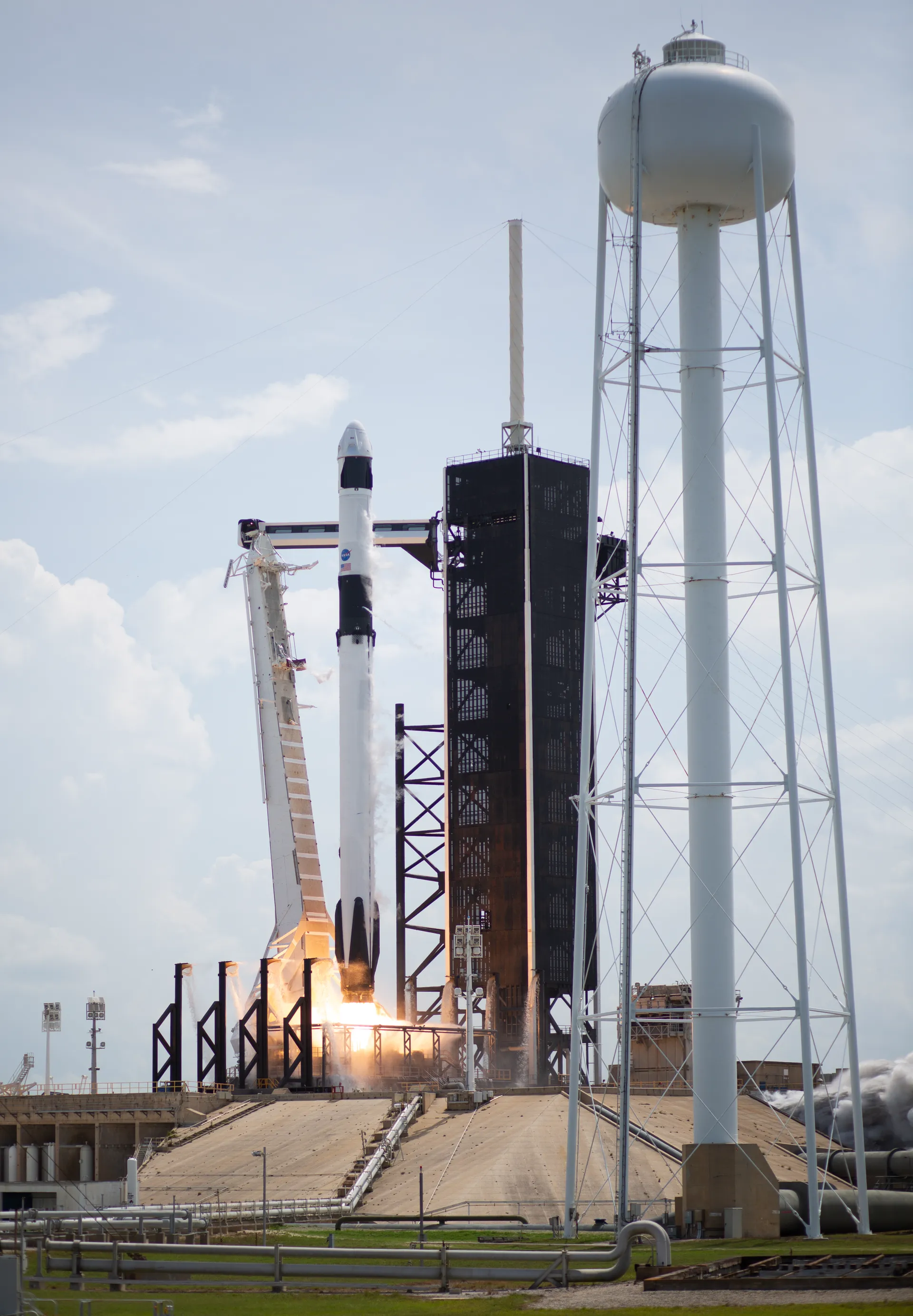 A Falcon 9 rocket lifts off beside its launch tower and a white water tower at Launch Complex 39A.