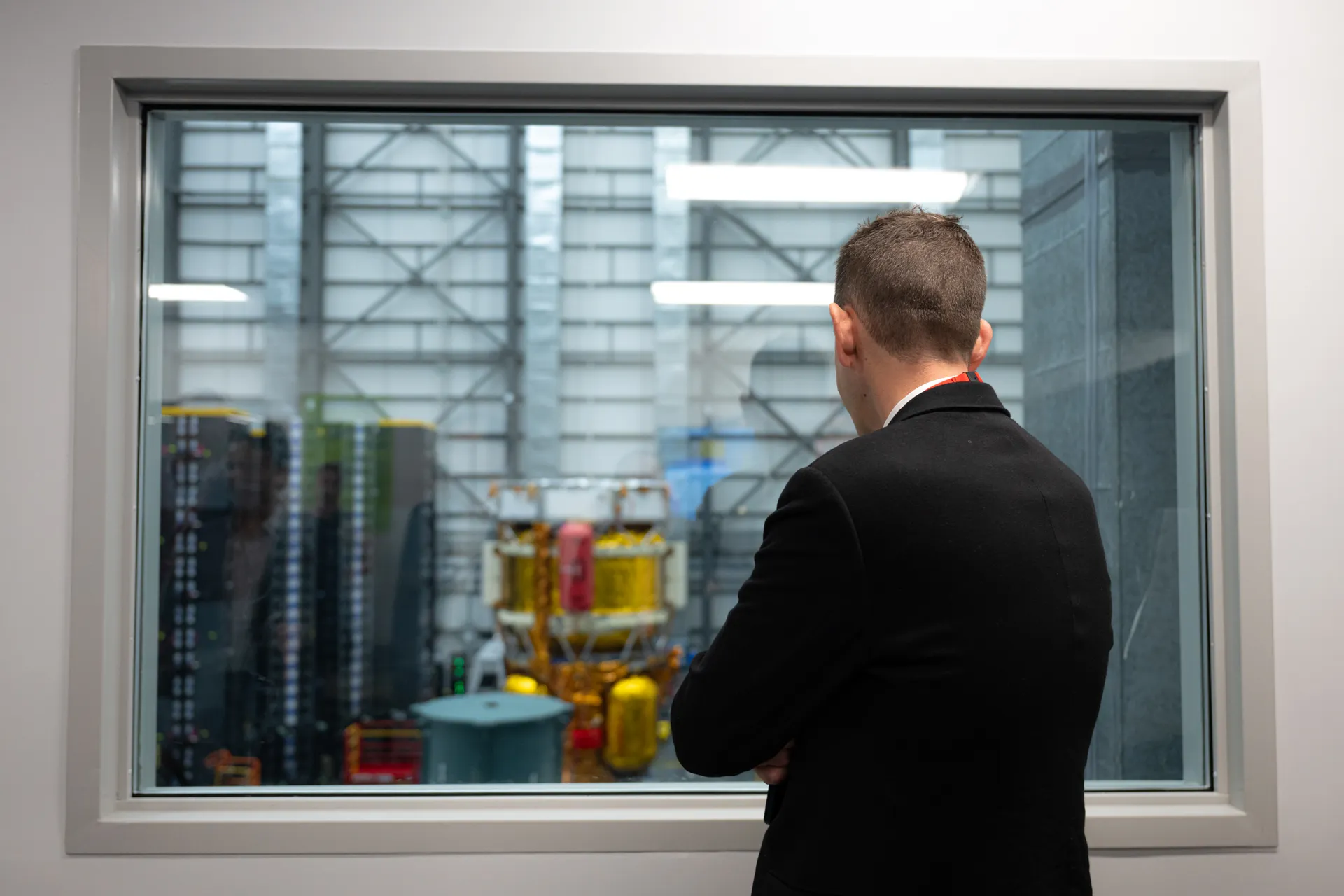 NASA Administrator Jared Isaacman, seen from behind, looks through a window at the gold-and-silver Blue Moon Mark 1 lander in Blue Origin’s factory.