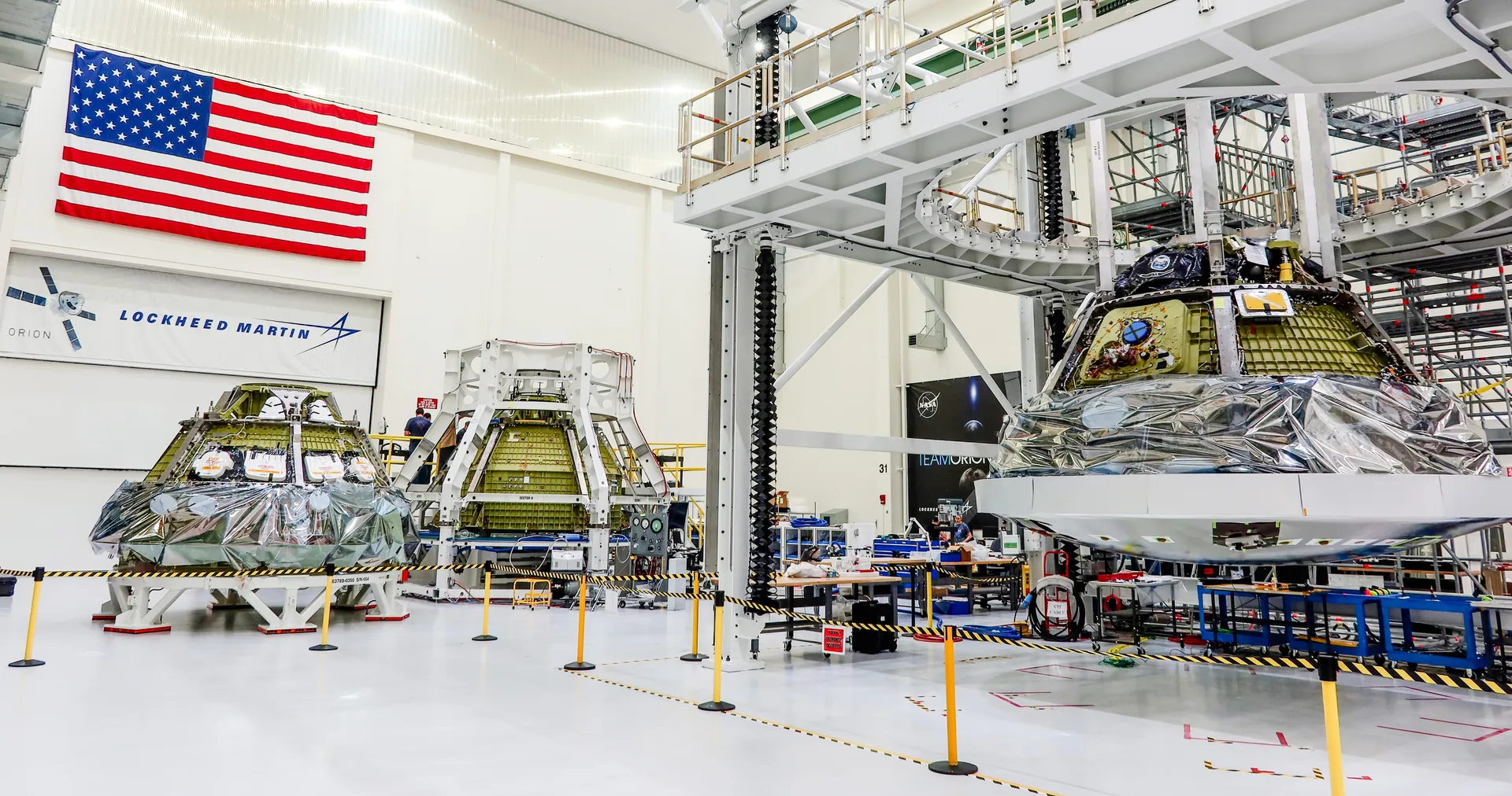 Three Orion crew modules in a bright high bay, one wrapped in silver foil, beside a Lockheed Martin banner.