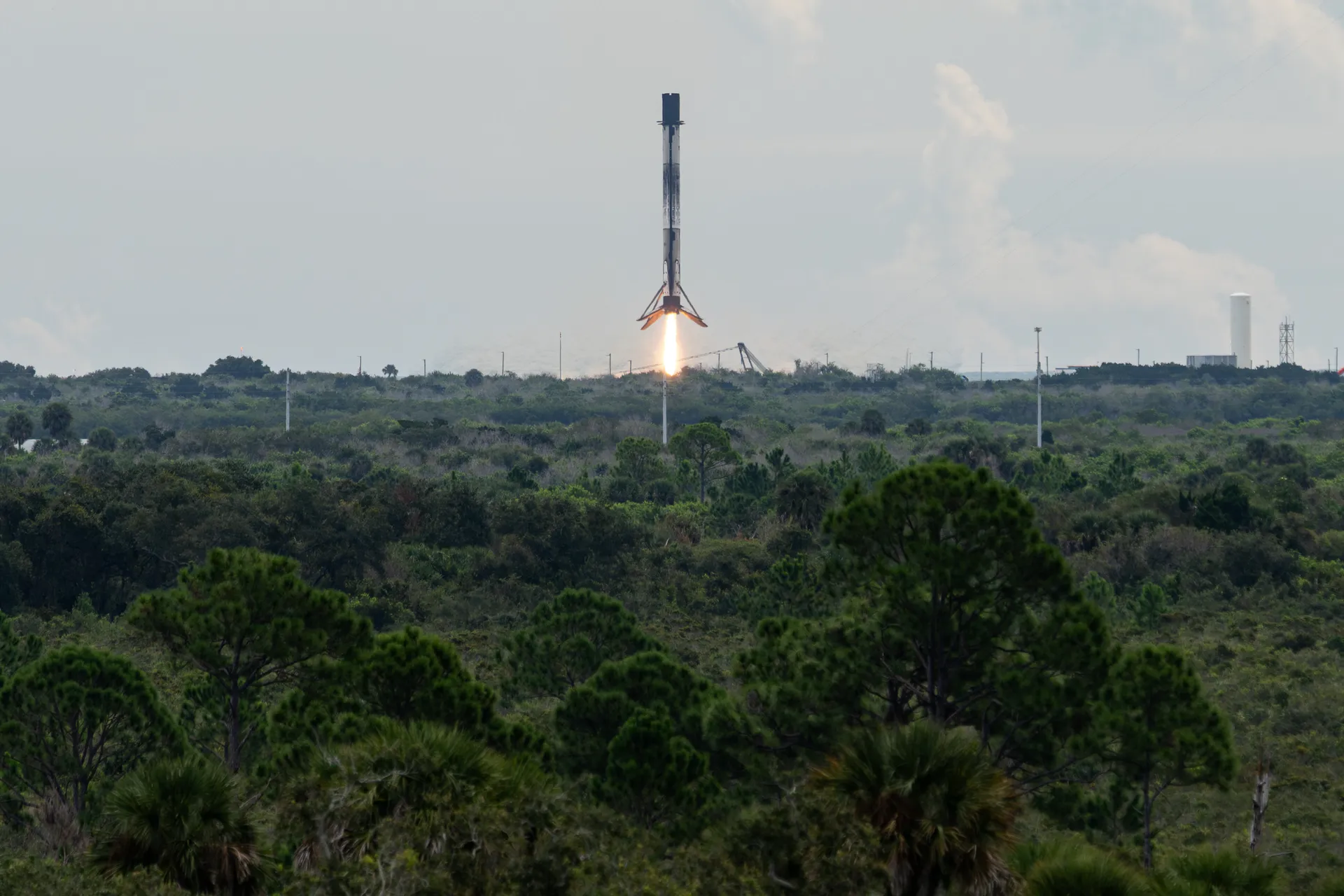 A Falcon 9 first stage, its legs spread, descends on a short flame toward a landing pad beyond a wide stretch of scrub forest.