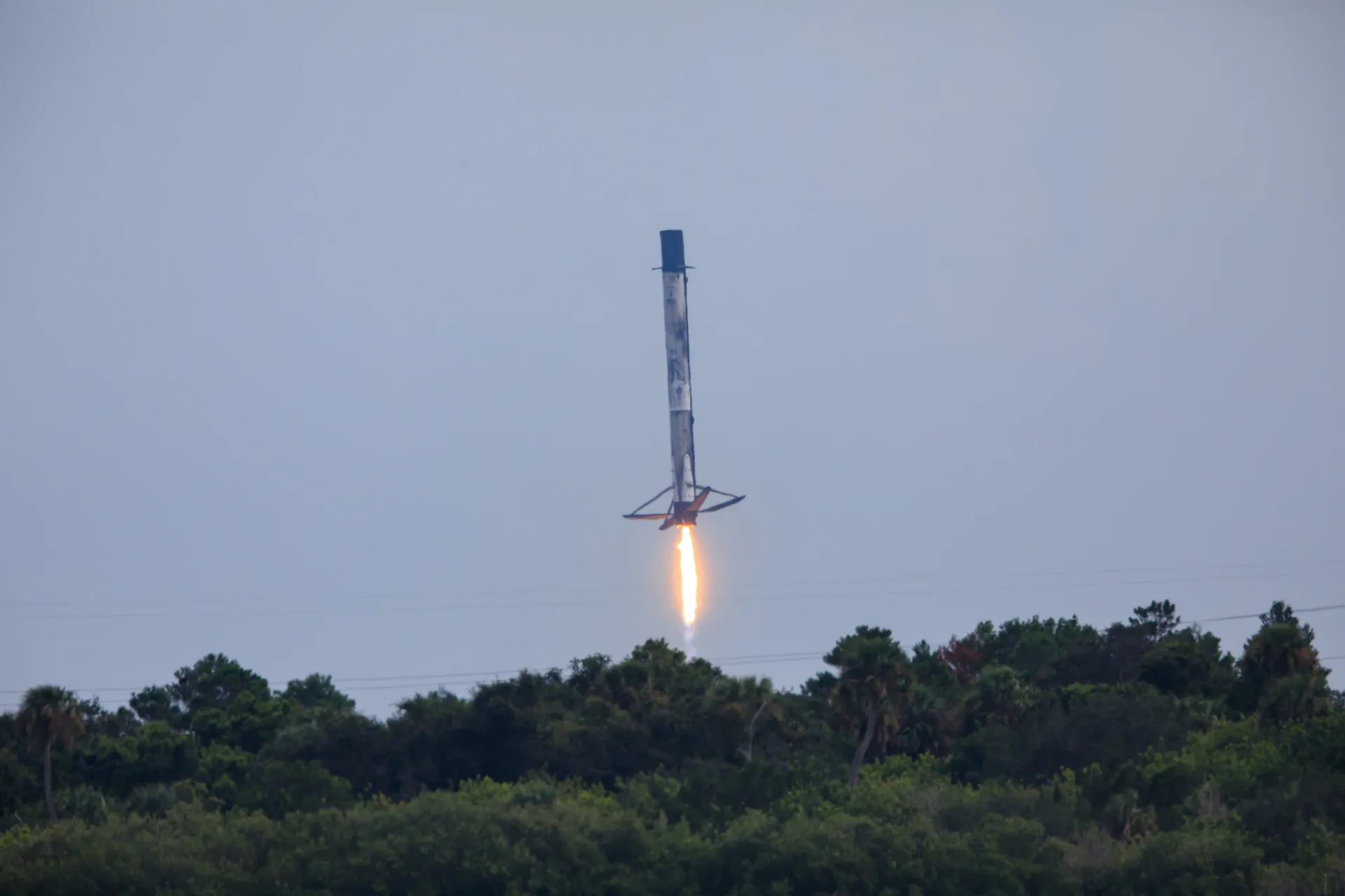 A Falcon 9 first stage descends on a single engine plume toward Landing Zone 1, its four legs deployed, above a line of trees.