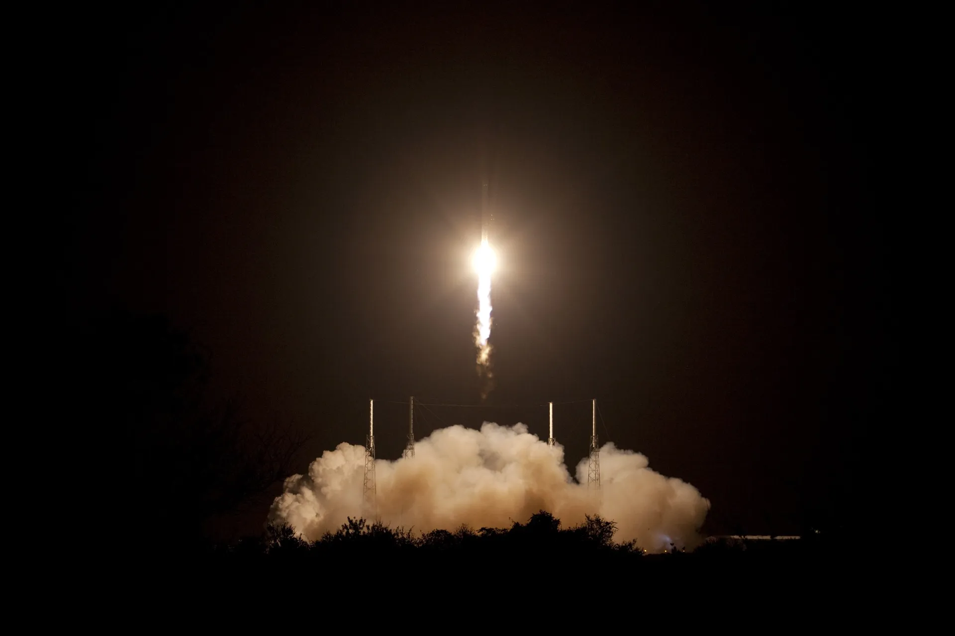 A Falcon 9 rises on a bright flame through the night sky over Space Launch Complex 40, lighting a cloud of exhaust on the ground.