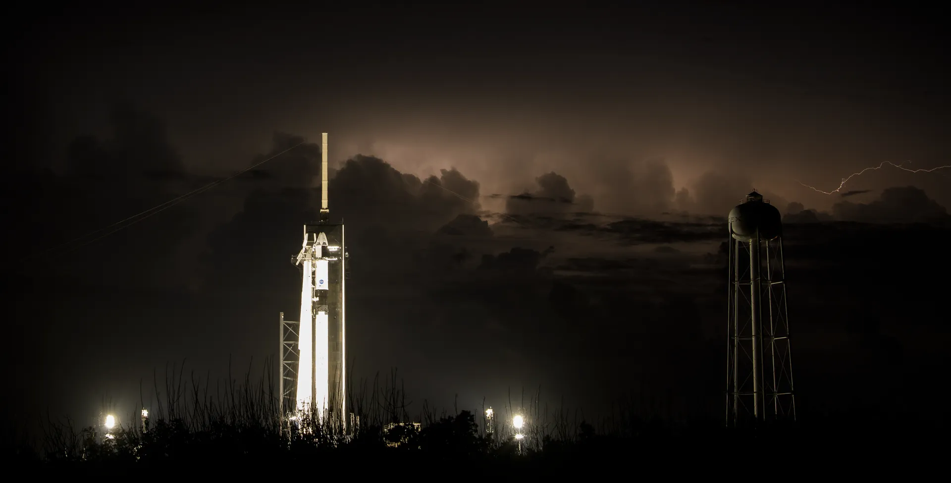 At night, a white Falcon 9 and Crew Dragon stand lit on Launch Complex 39A as lightning flickers in distant storm clouds.