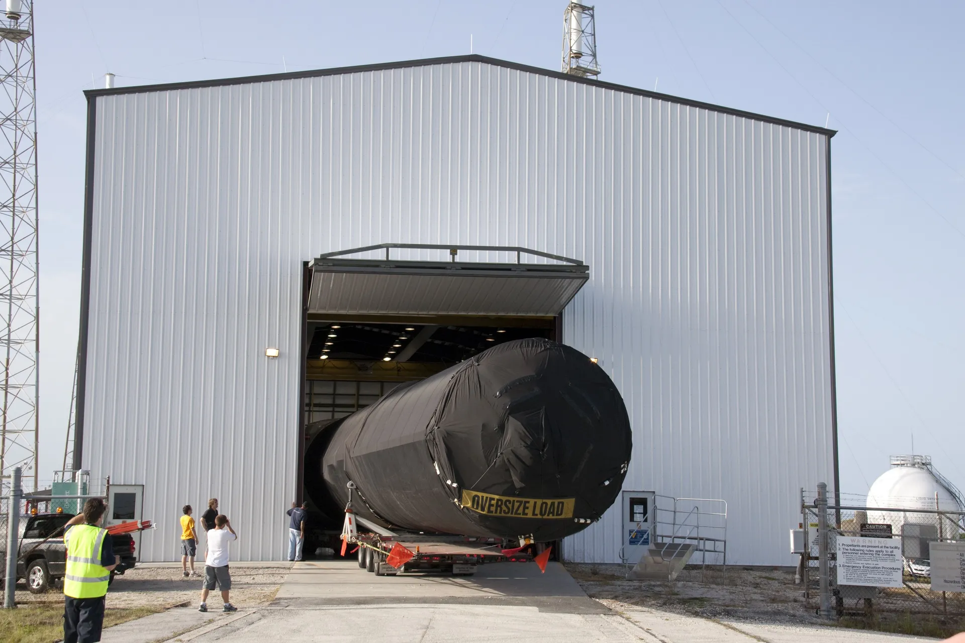 A Falcon 9 first stage under a black protective cover is backed on a trailer into a corrugated-metal hangar at Space Launch Complex 40.