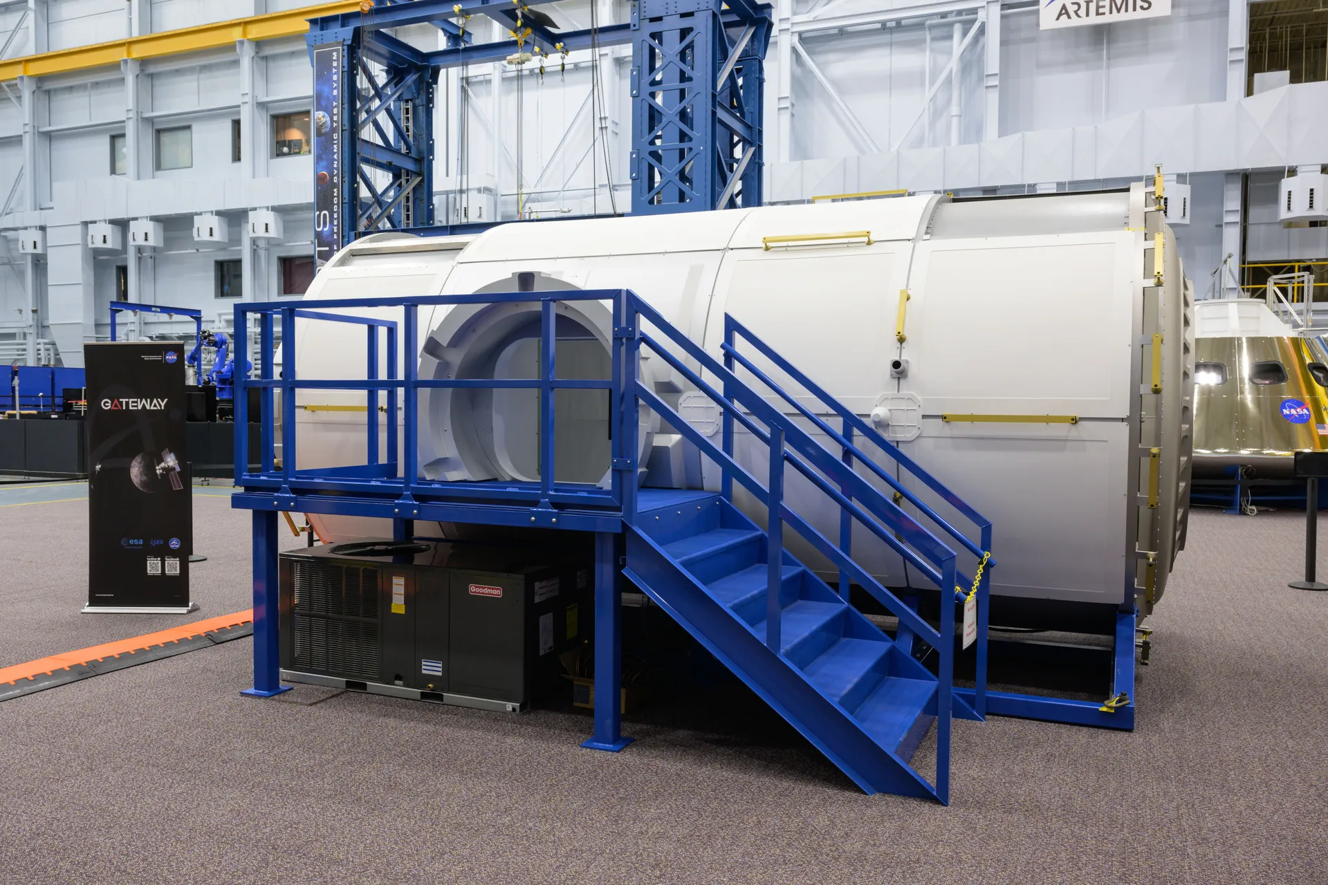 A full-size white mockup of the HALO module, with blue stairs leading up to its open hatch, stands in a large training hall beside an Orion mockup.