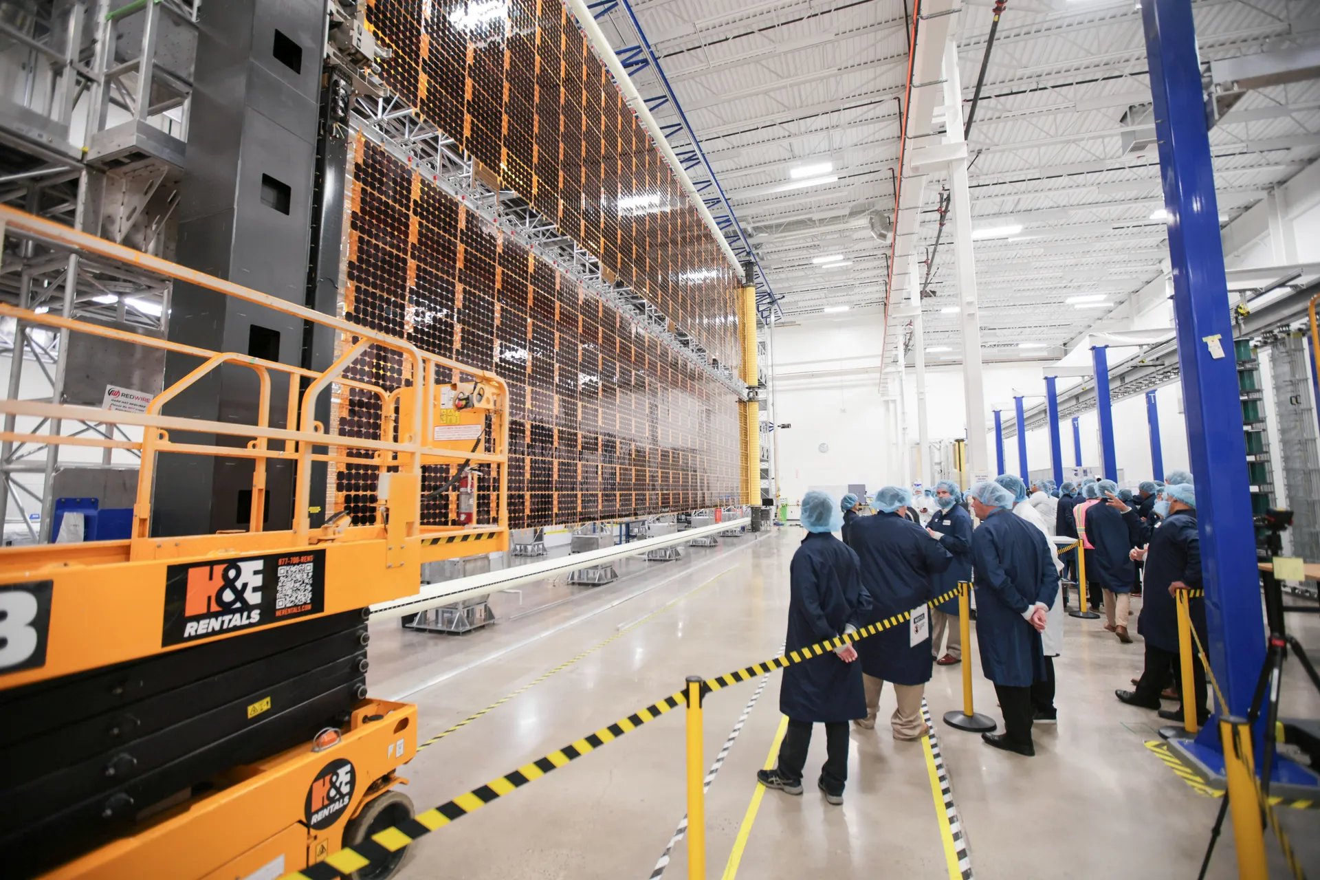 A tall Roll-Out Solar Array wing stands fully extended along one side of a long, brightly lit high bay while visitors in hairnets and lab coats watch from behind a barrier.