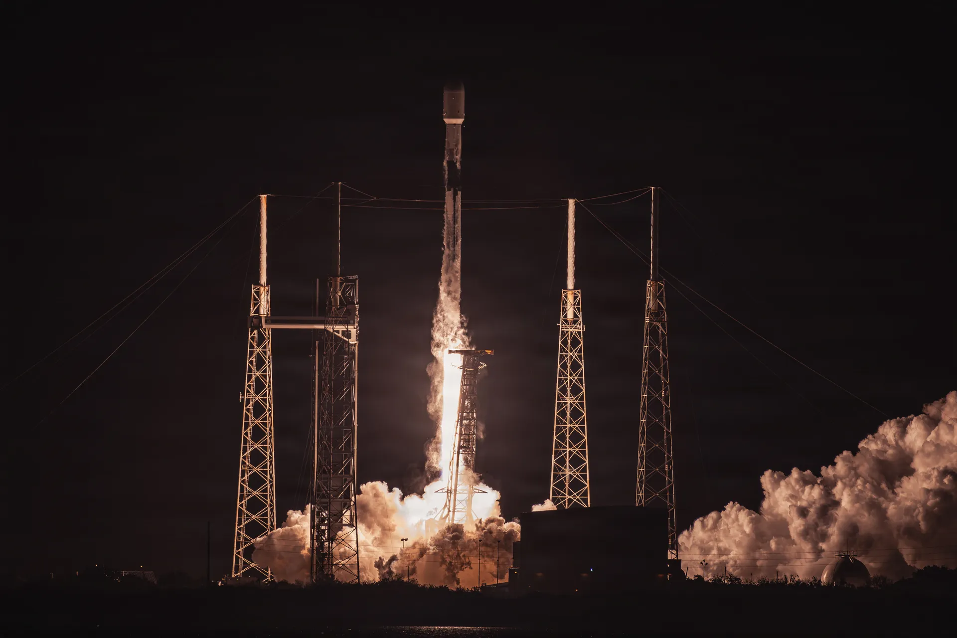A Falcon 9 climbs on a column of flame between the lit lightning towers of Space Launch Complex 40 at night, with exhaust clouds rolling across the pad.