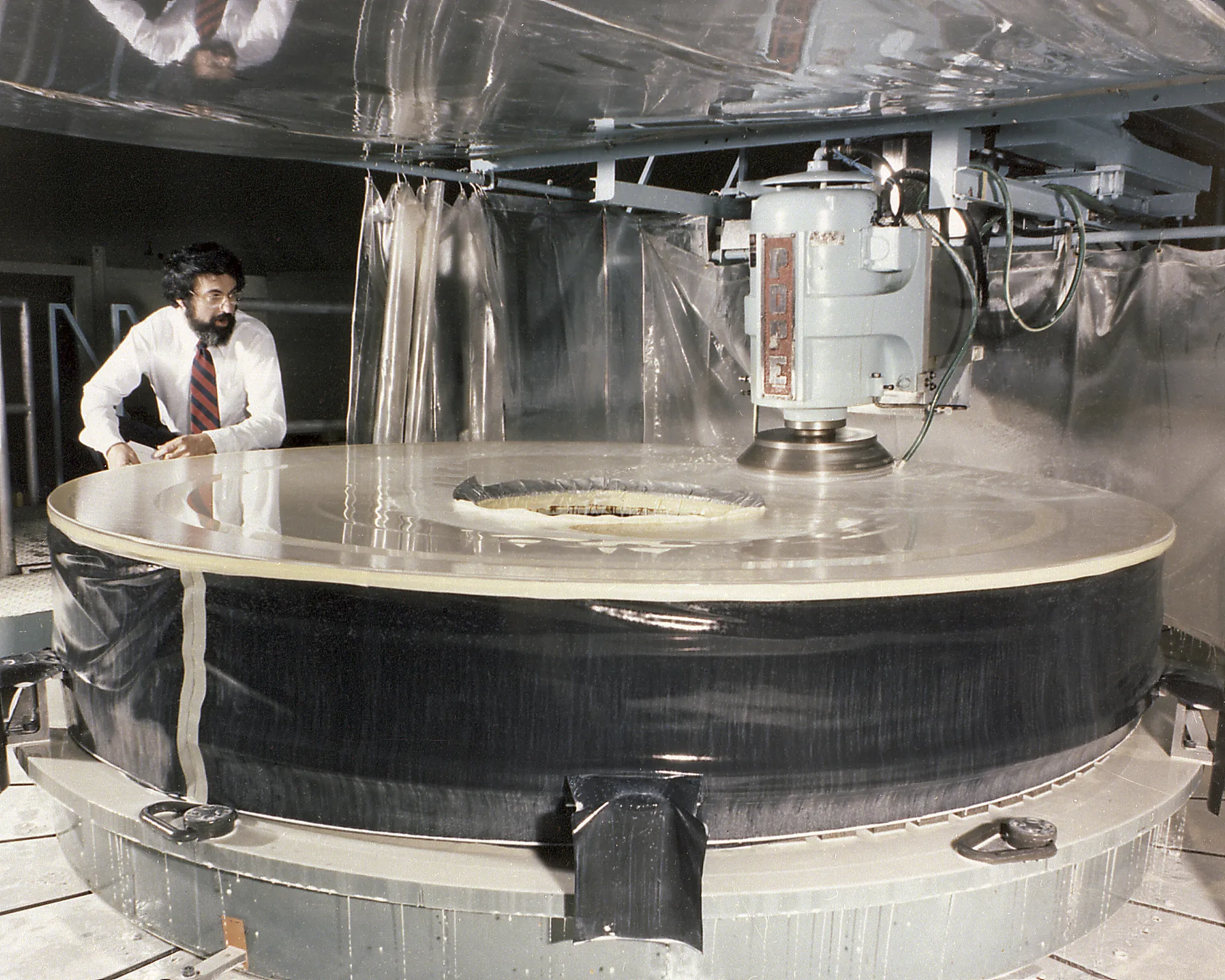 An engineer in a white shirt and tie stands beside a huge round glass blank on a grinding machine in a factory clean area.