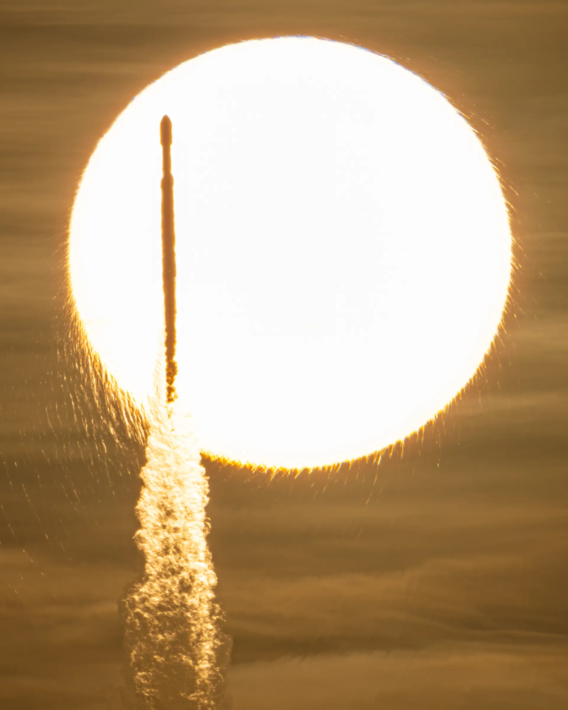 Silhouetted against a huge orange disk of the Sun, a rocket climbs on a pillar of exhaust.
