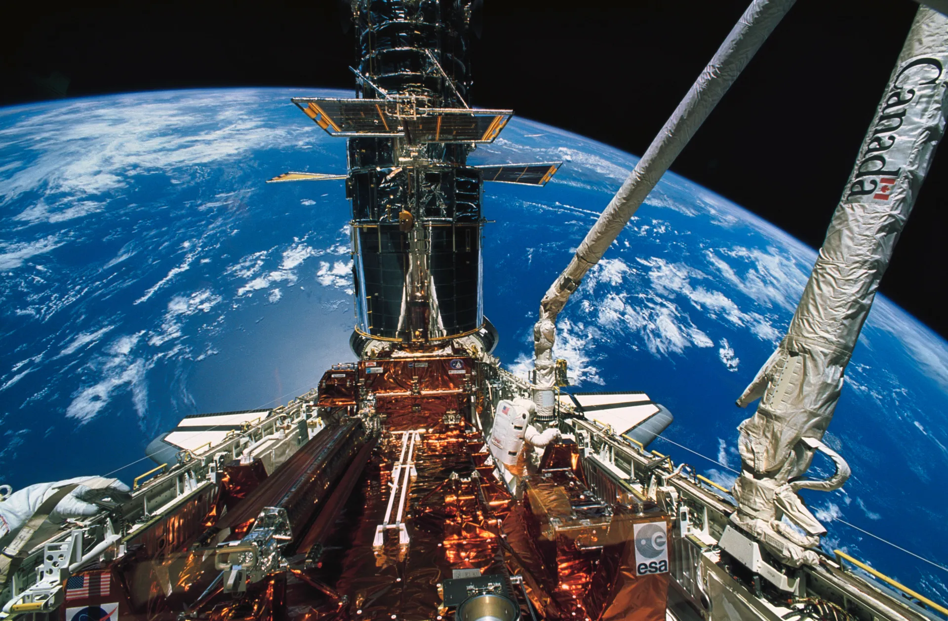 Hubble stands upright in the Space Shuttle Endeavour’s cargo bay above the blue Earth, with the robotic arm and an astronaut beside it.