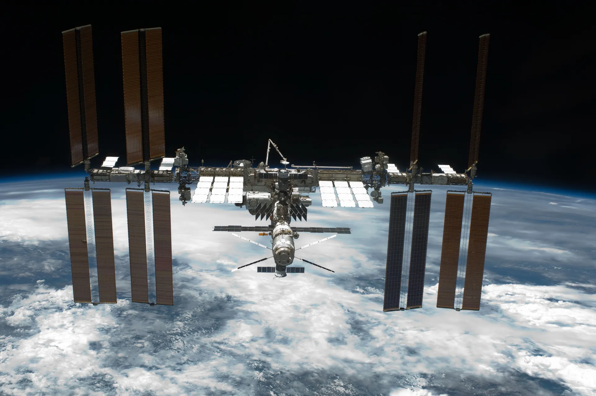 The complete station with eight long solar-array wings above a cloudy Earth.