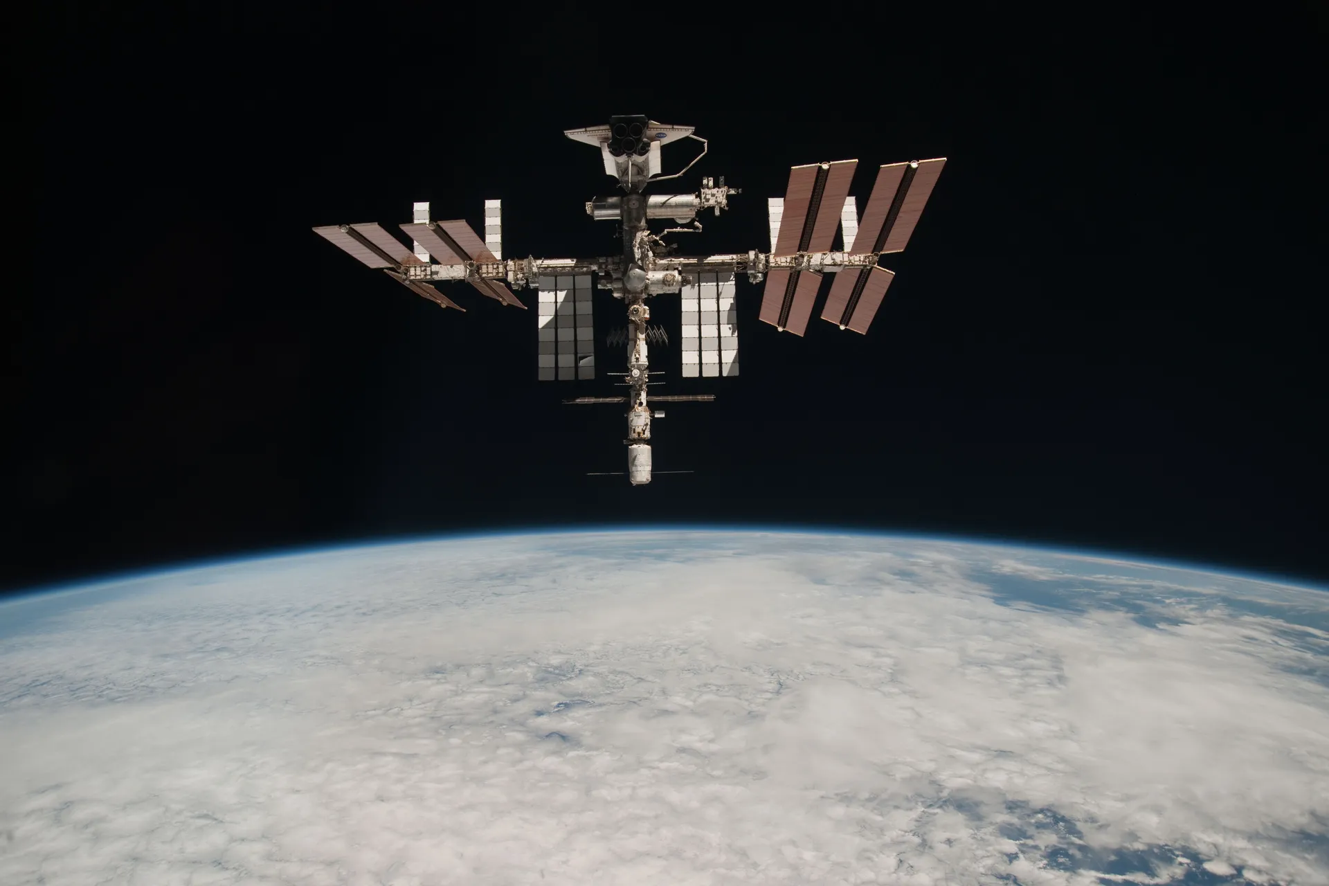 The completed International Space Station with the space shuttle Endeavour docked, its eight solar-array wings spread above the cloud-covered curve of Earth.
