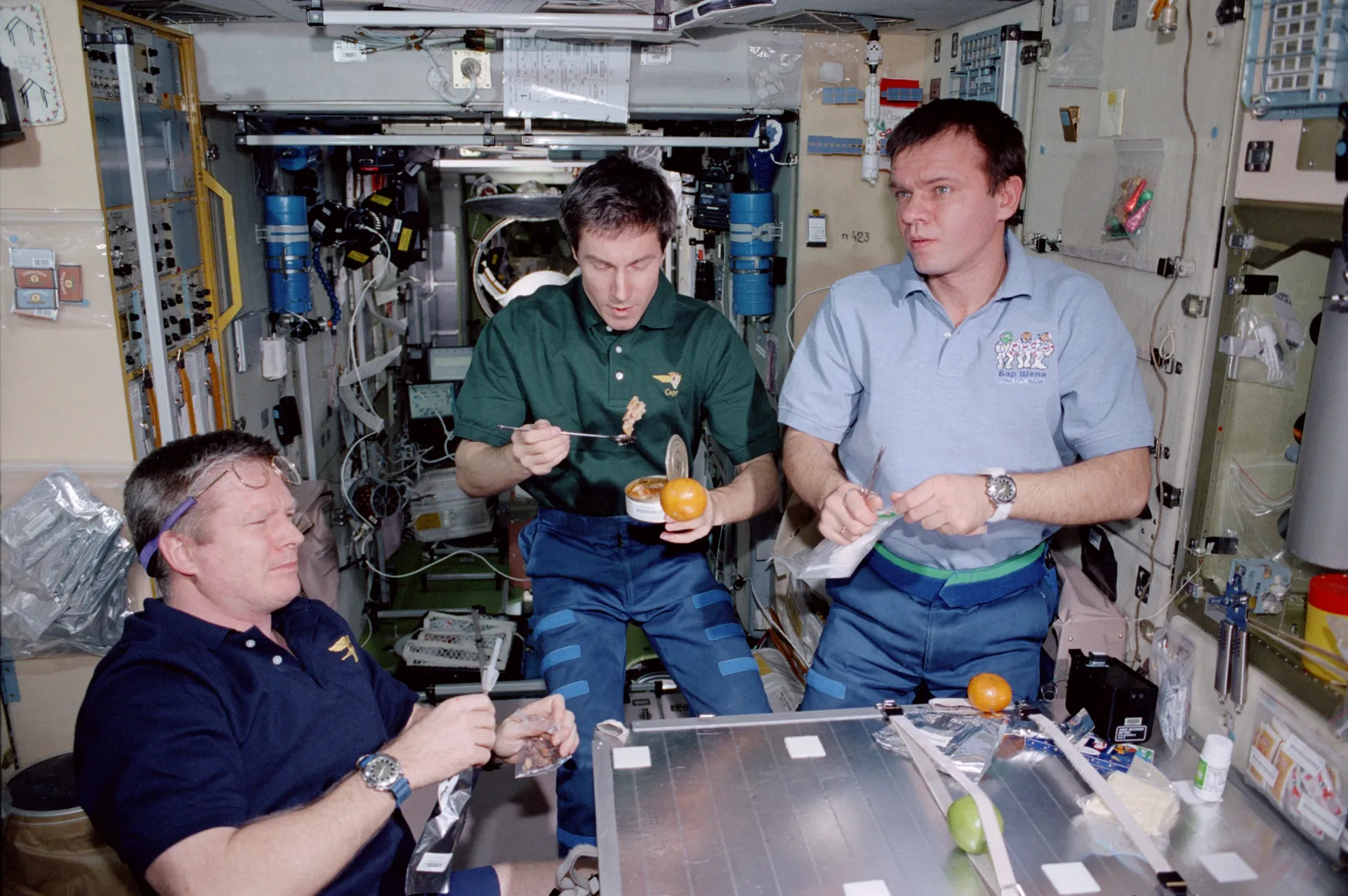 Three crew members in casual clothes float around a small table inside a cluttered module, holding food.