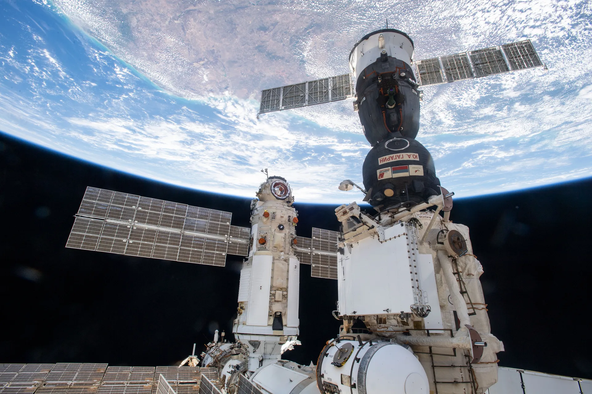 A large white Russian module and a docked Soyuz spacecraft above the curve of Earth.