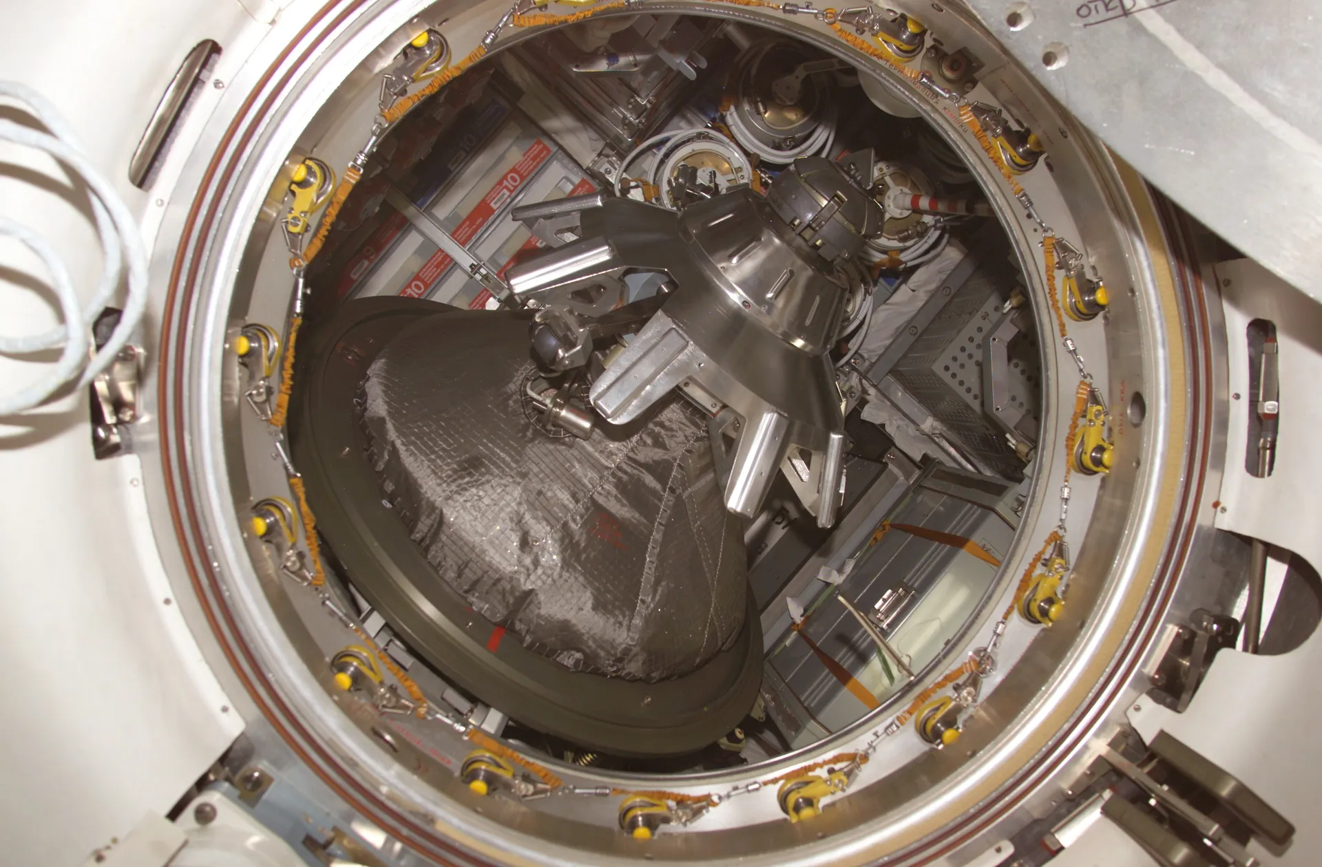A round open hatch ringed with latches, looking into a docked spacecraft with a cone-shaped docking mechanism.