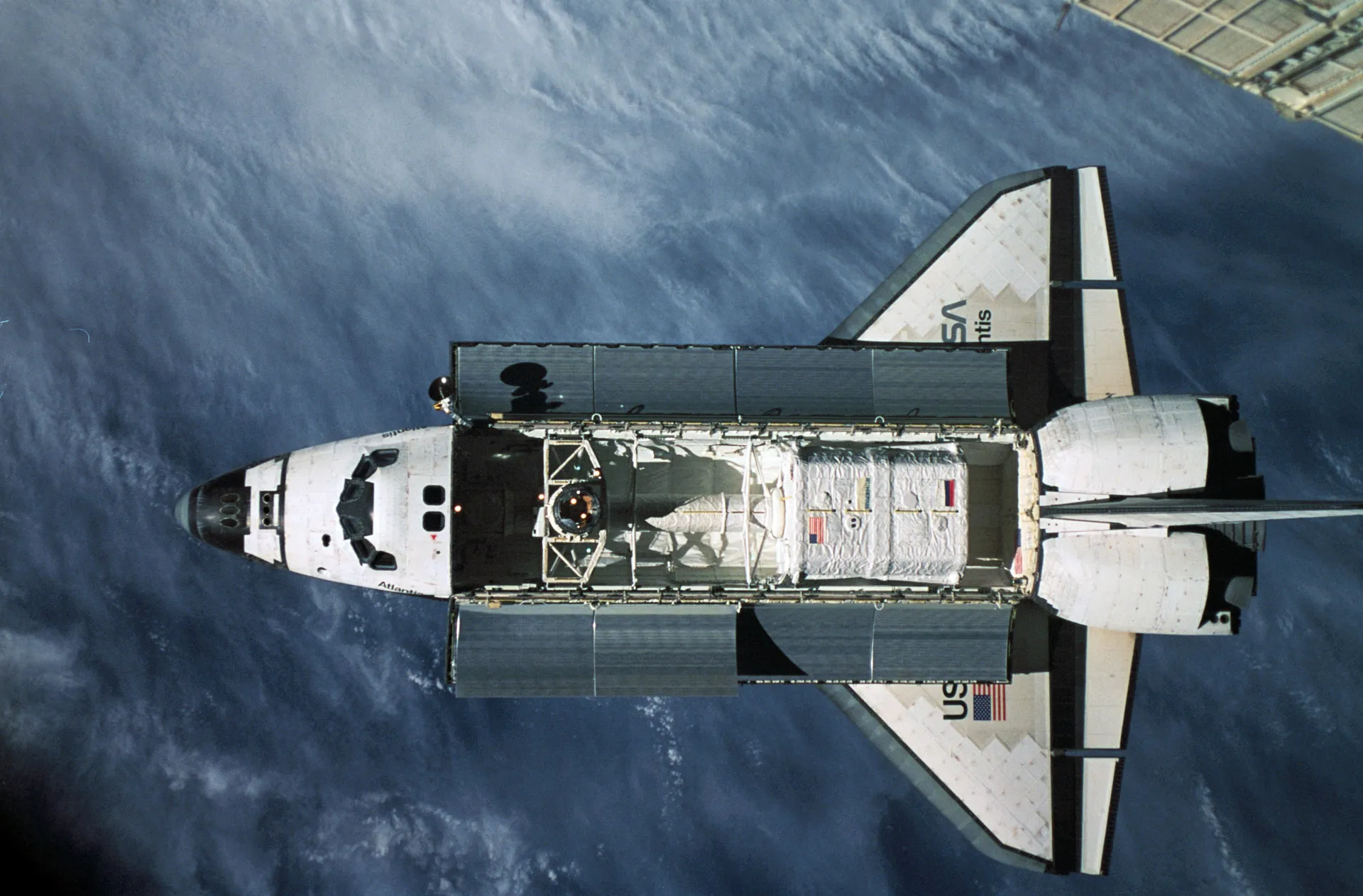 The space shuttle Atlantis seen from above against clouds and ocean, its payload bay open, approaching the photographer.