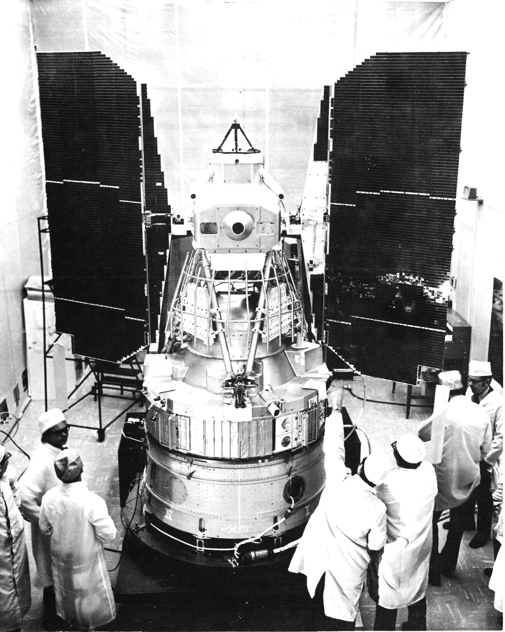 Black-and-white photograph of the ERTS-1 spacecraft in a high bay: a tall cylindrical body with two large solar panels raised like wings, surrounded by engineers in white coats and caps.