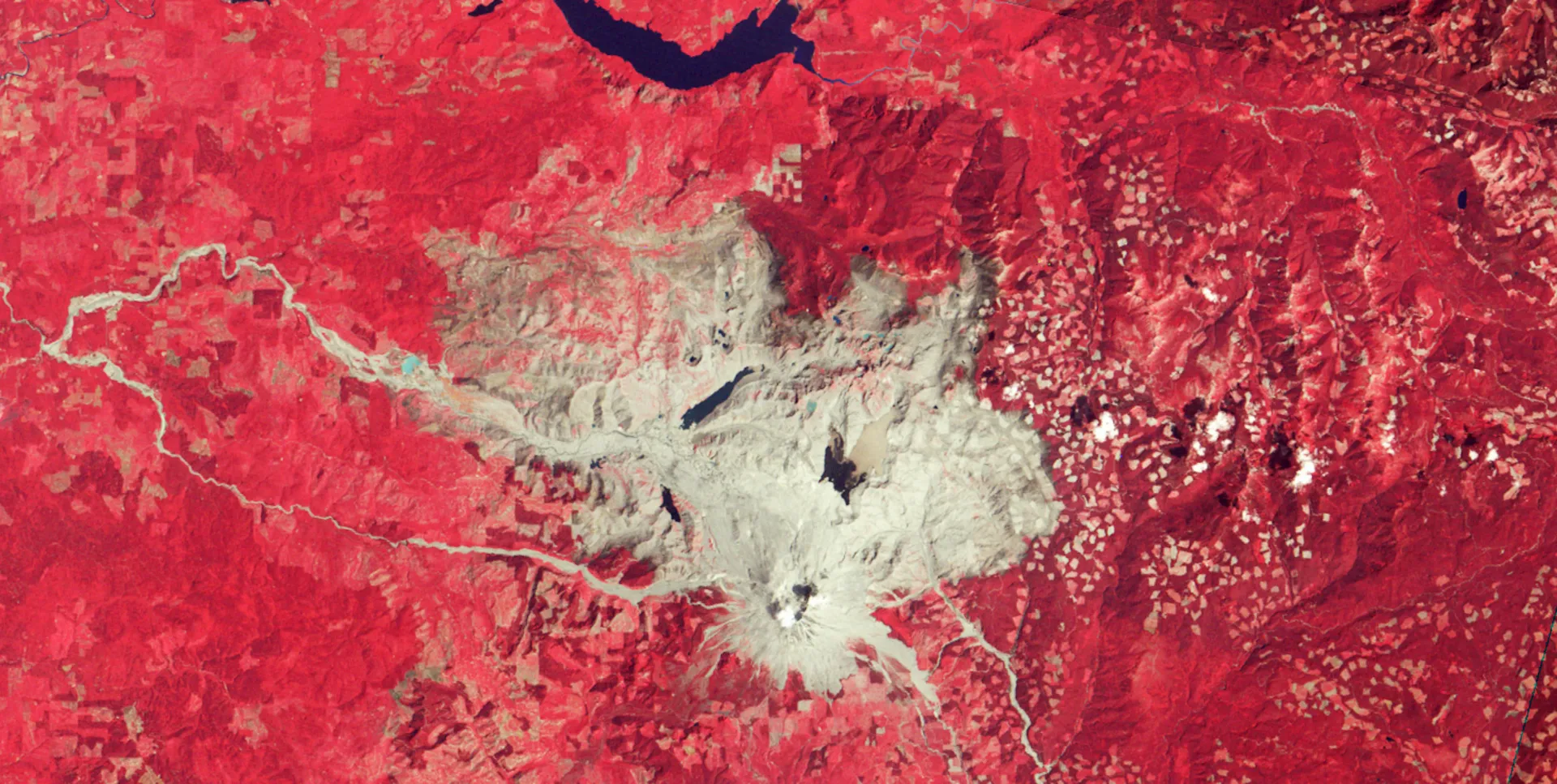 The same area after the eruption: a broad gray fan of ash and rock spreads north of the volcano through the red forest.