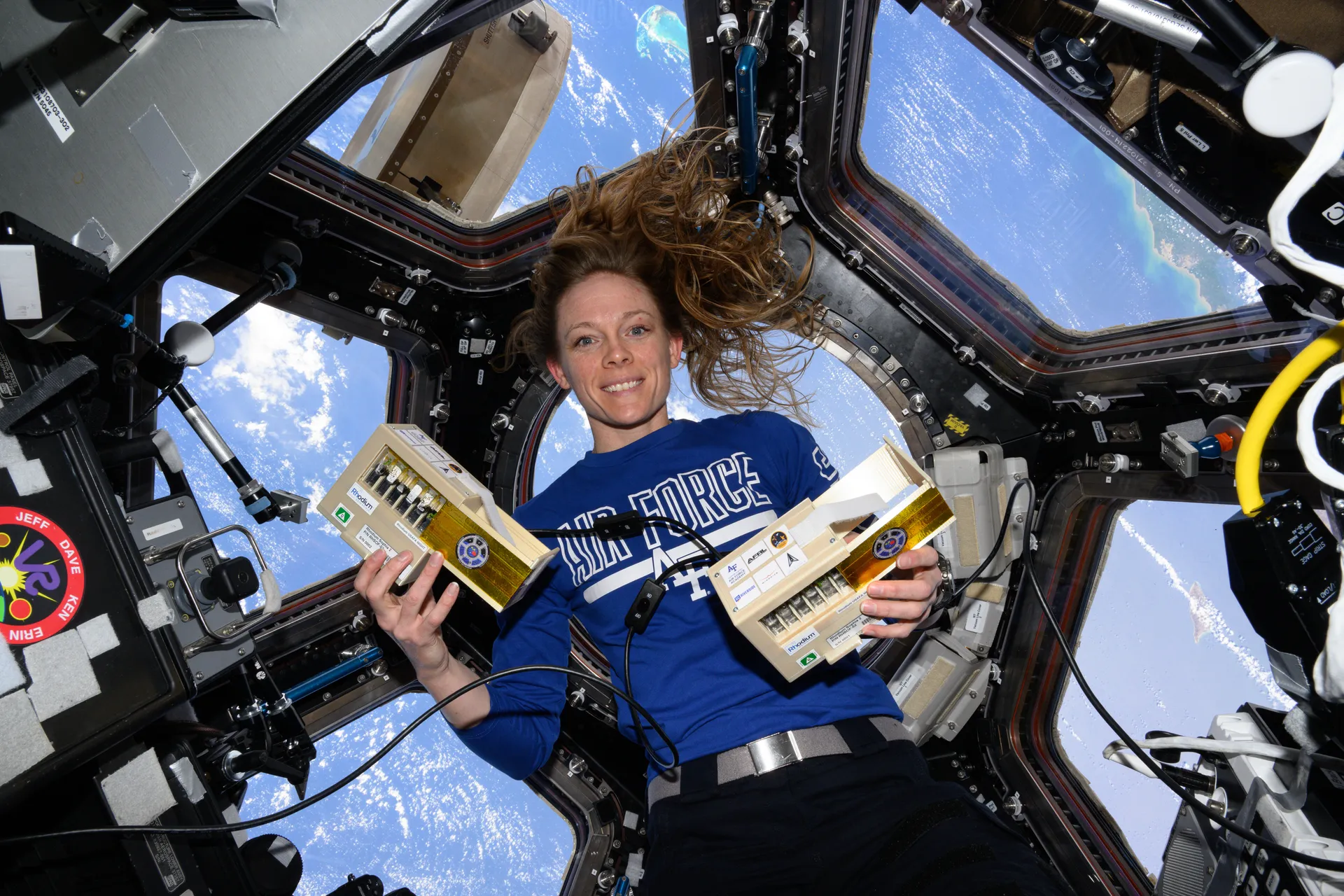 Nichole Ayers floats in the cupola holding two small plant-growth boxes, with Earth’s ocean and clouds visible through the windows around her.