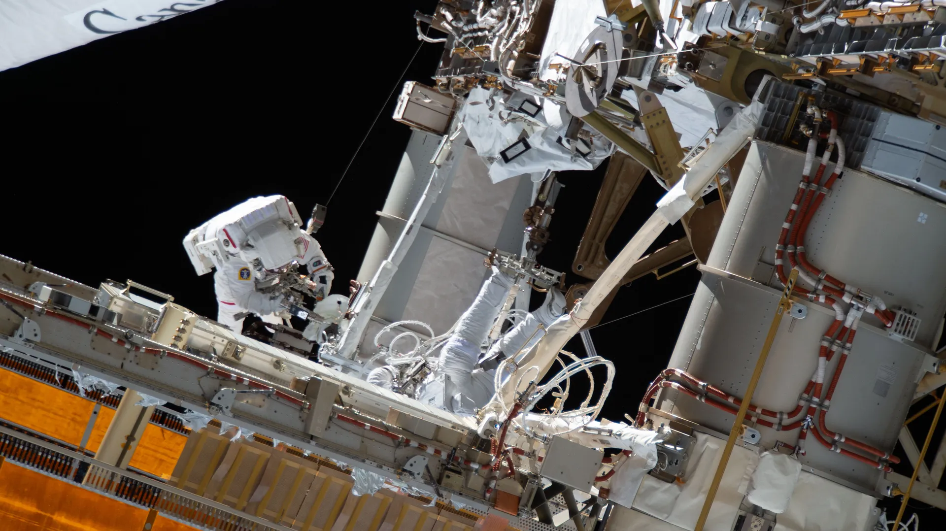 Jessica Meir in a white spacesuit works on the station’s truss against black space, beside a tangle of cables, boxes and the gold edge of a solar array.