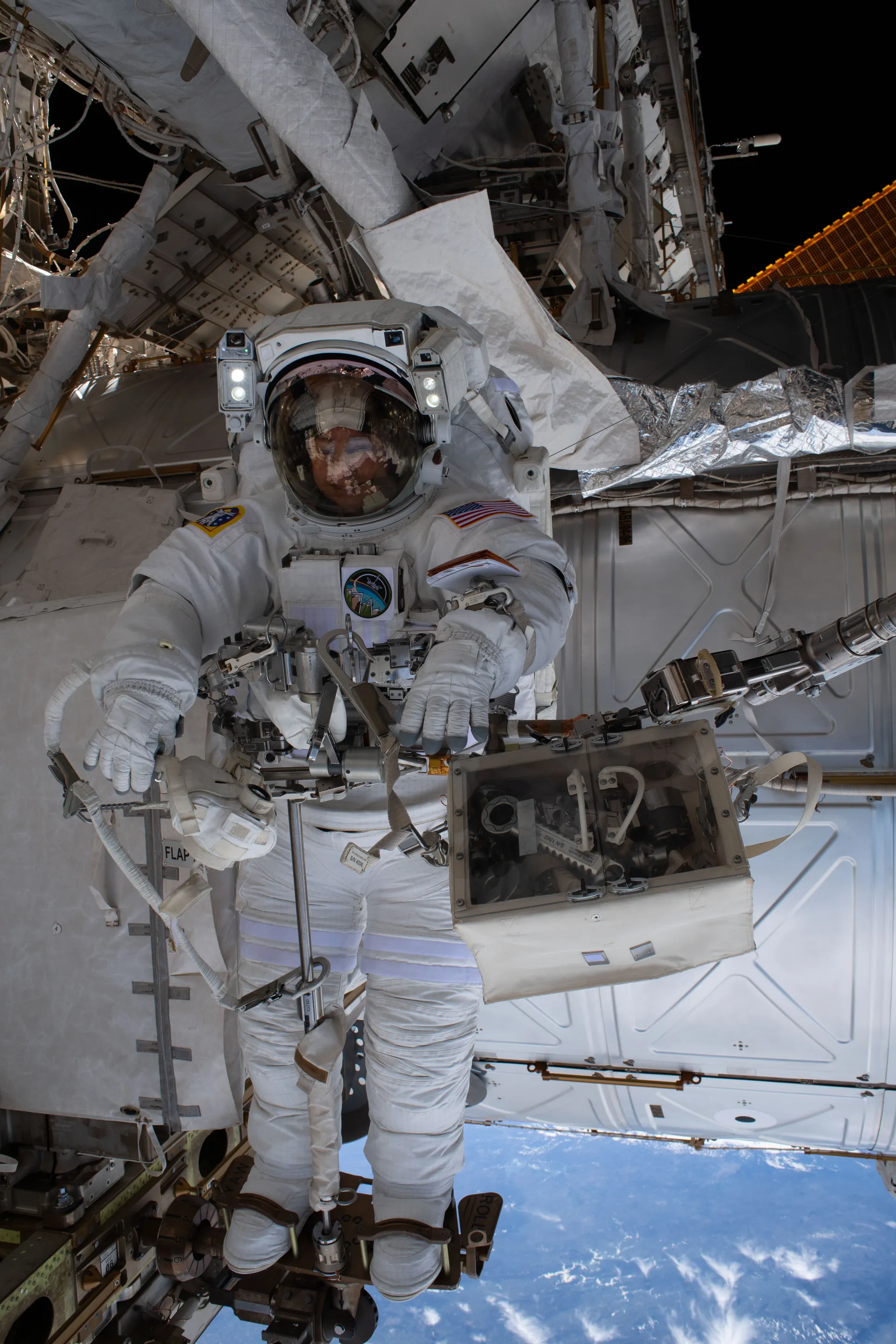Jessica Meir in a white spacesuit stands upright in a foot restraint on the end of the station’s robotic arm, with the blue Earth far below.