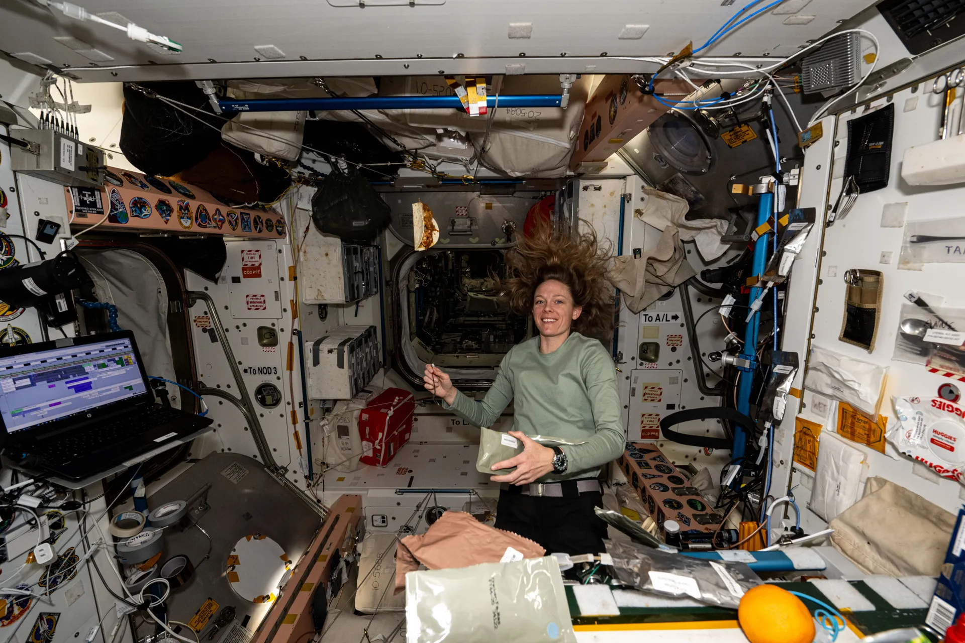 Nichole Ayers floats beside the galley in the Unity module, holding a food packet, with a laptop and food containers fastened around her.