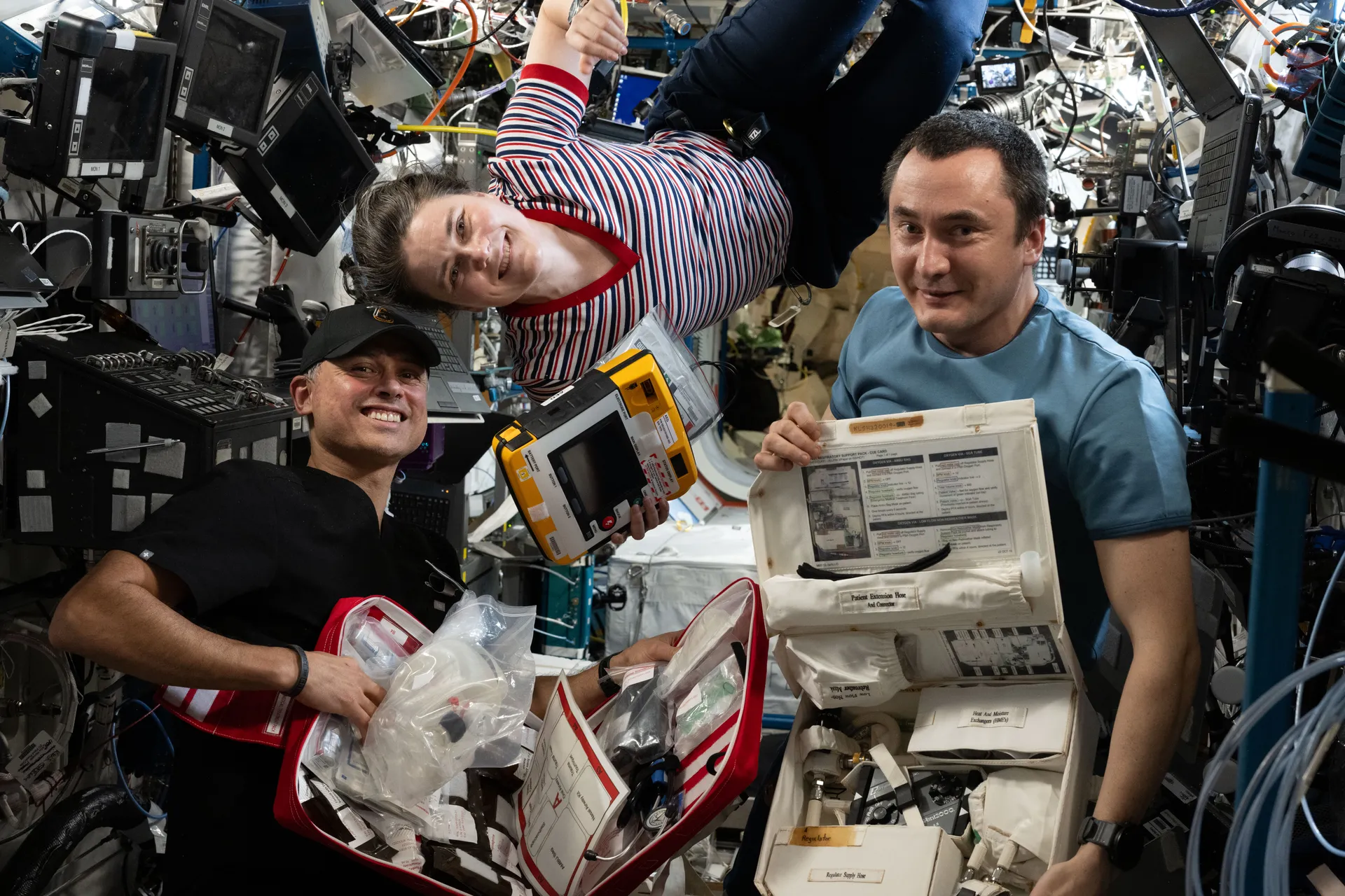 Anil Menon, Anna Kikina and Pyotr Dubrov hold open emergency medical kits and a defibrillator in the Destiny laboratory.