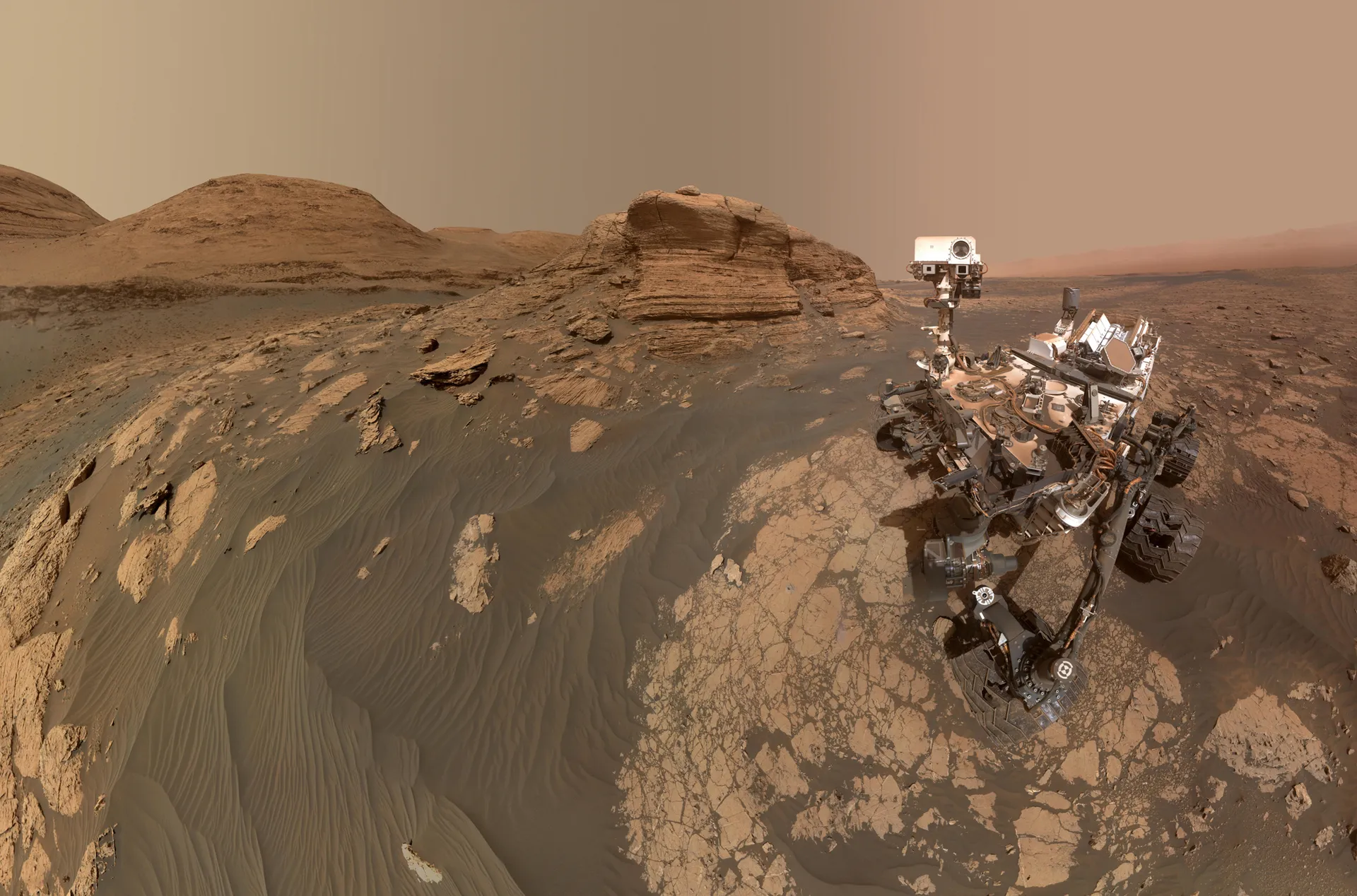 Curiosity, a car-sized rover with a tall mast, sits on pale rock in front of a layered sandstone cliff under a tan sky.