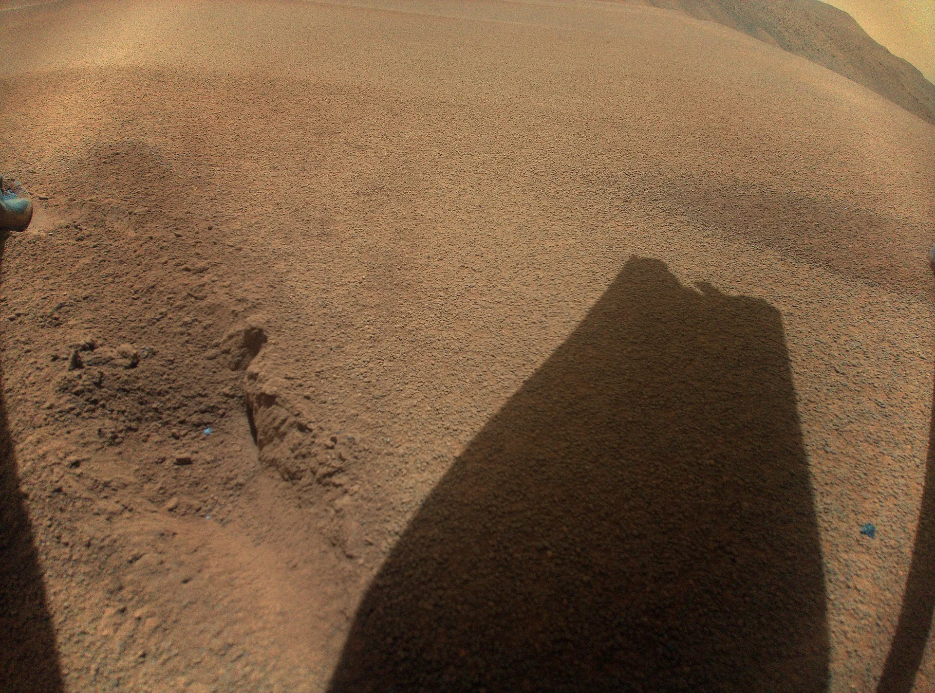 A fisheye view of tan sand with the dark shadow of a broken rotor blade across it.