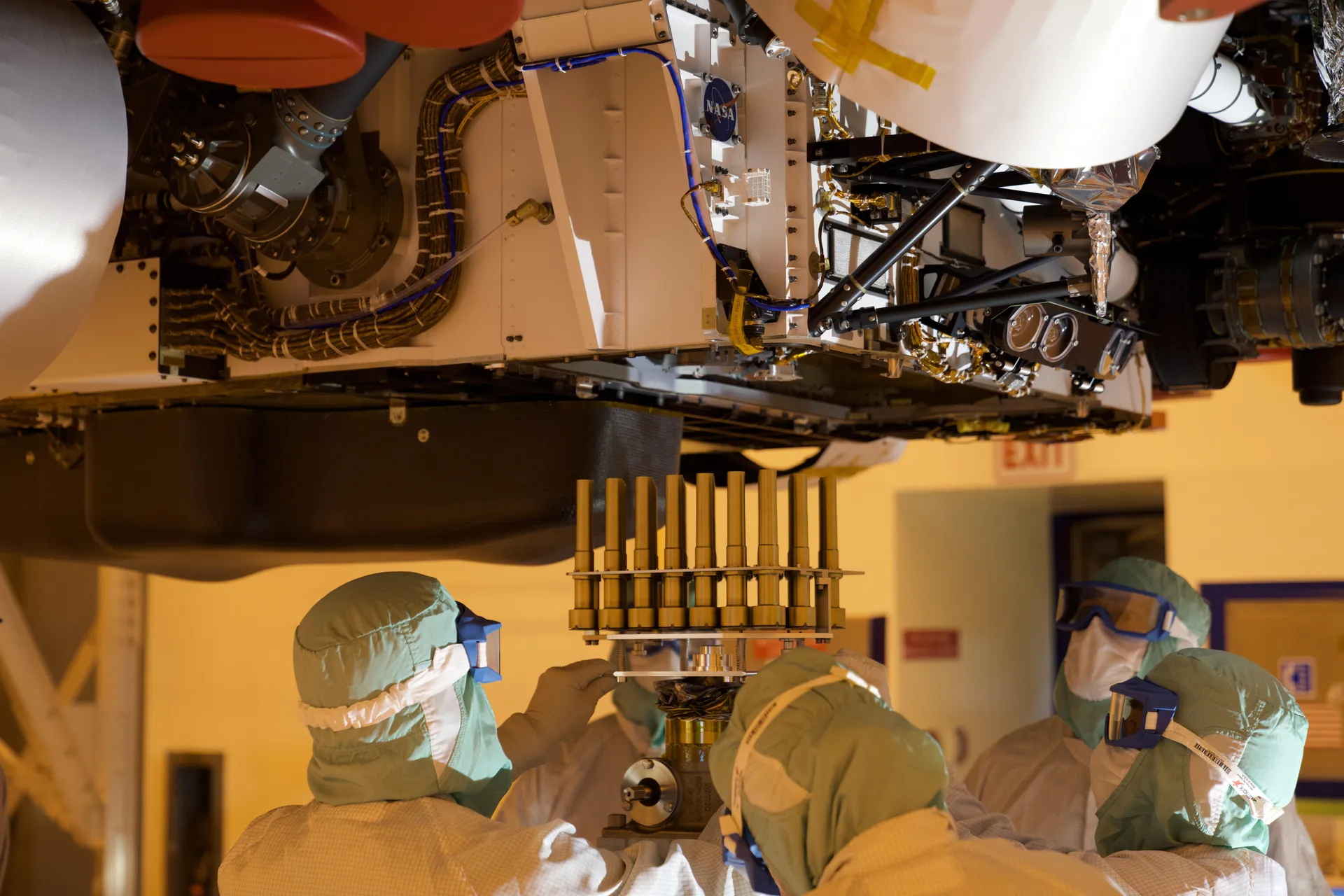 Technicians in full clean-room suits and goggles fit a tray of gold-sheathed tubes into the underside of the rover in a yellow-lit clean room.