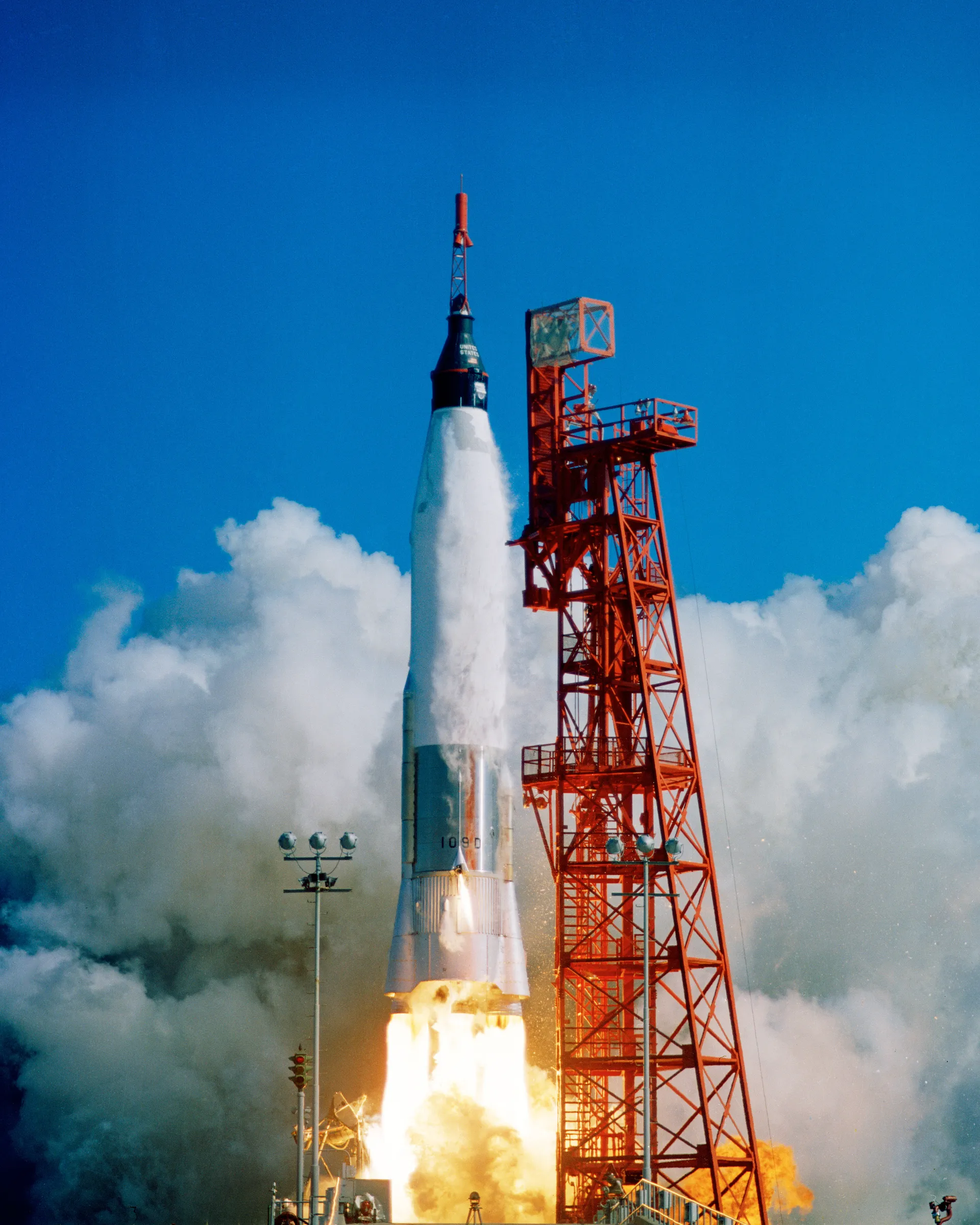 A silver Mercury-Atlas rocket climbs past its red service tower in billowing smoke.