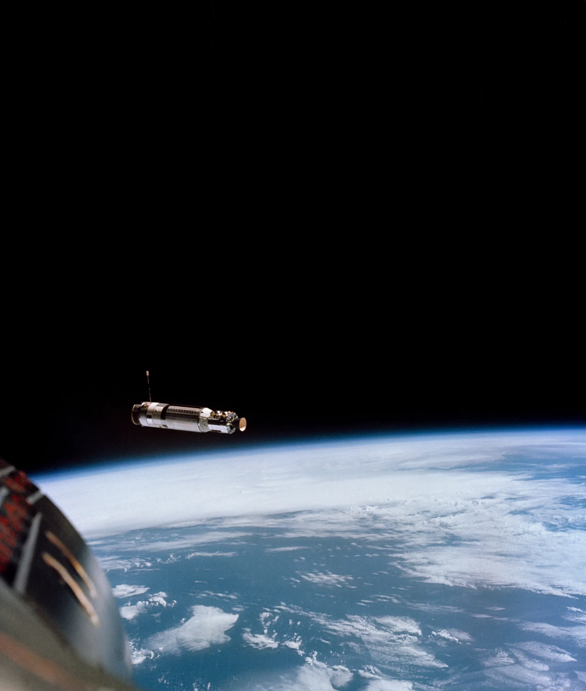 The small Agena target vehicle against black space above Earth’s blue edge, with the nose of Gemini 8 in the corner.
