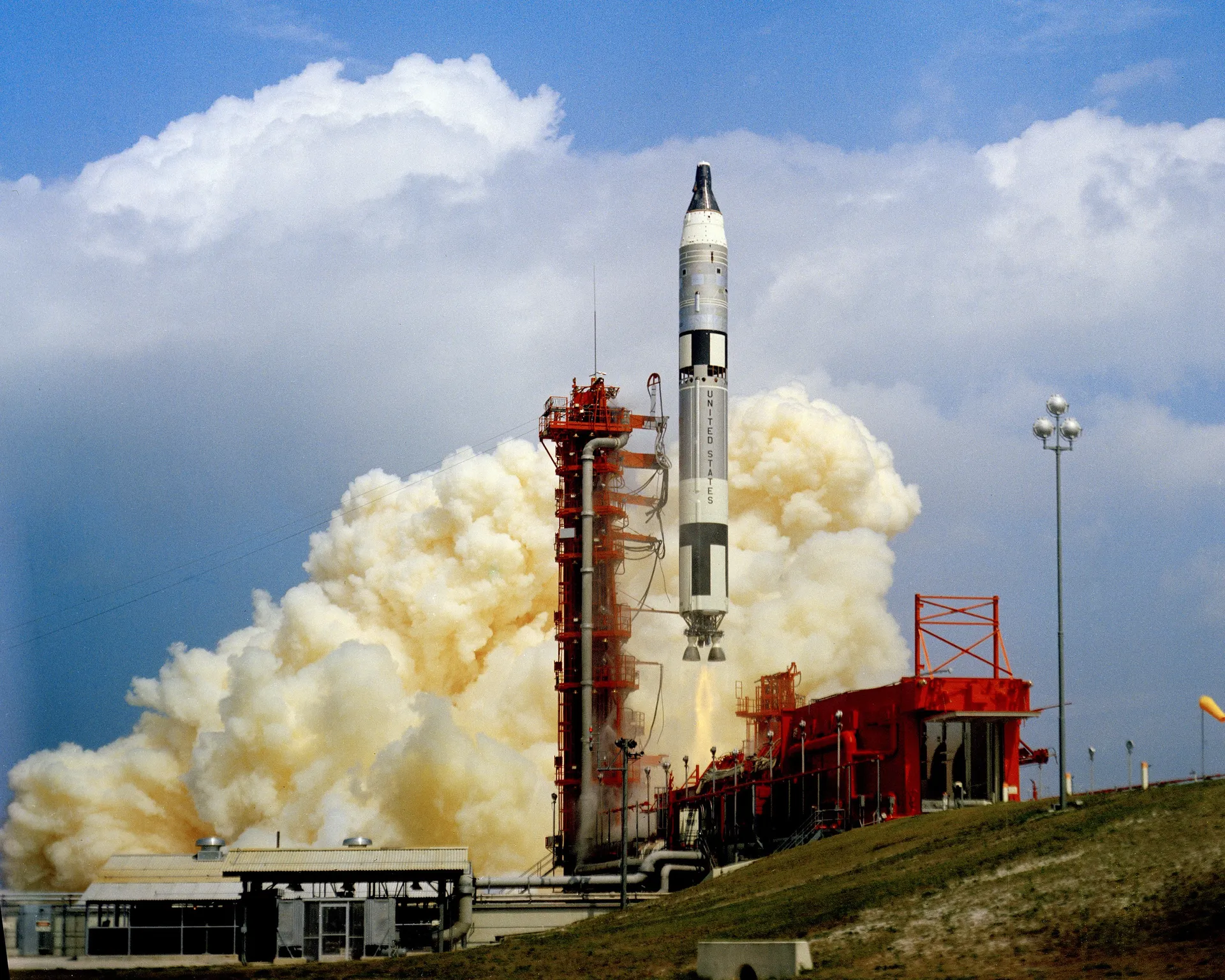 A Titan II rocket climbs from Launch Complex 19 beside its red erector, trailing smoke.