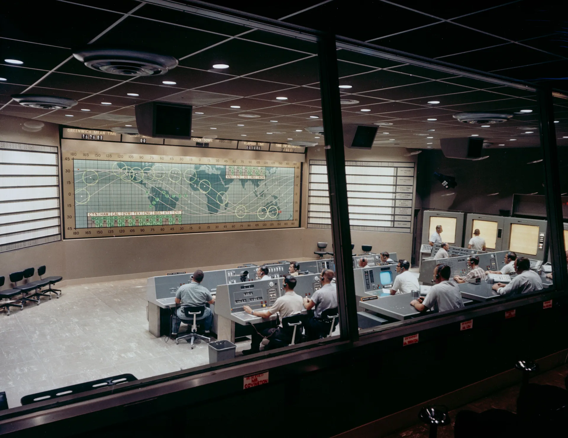 Flight controllers at rows of consoles in the Mercury Control Center at Cape Canaveral, facing a wall-size world map with the capsule’s orbital track.