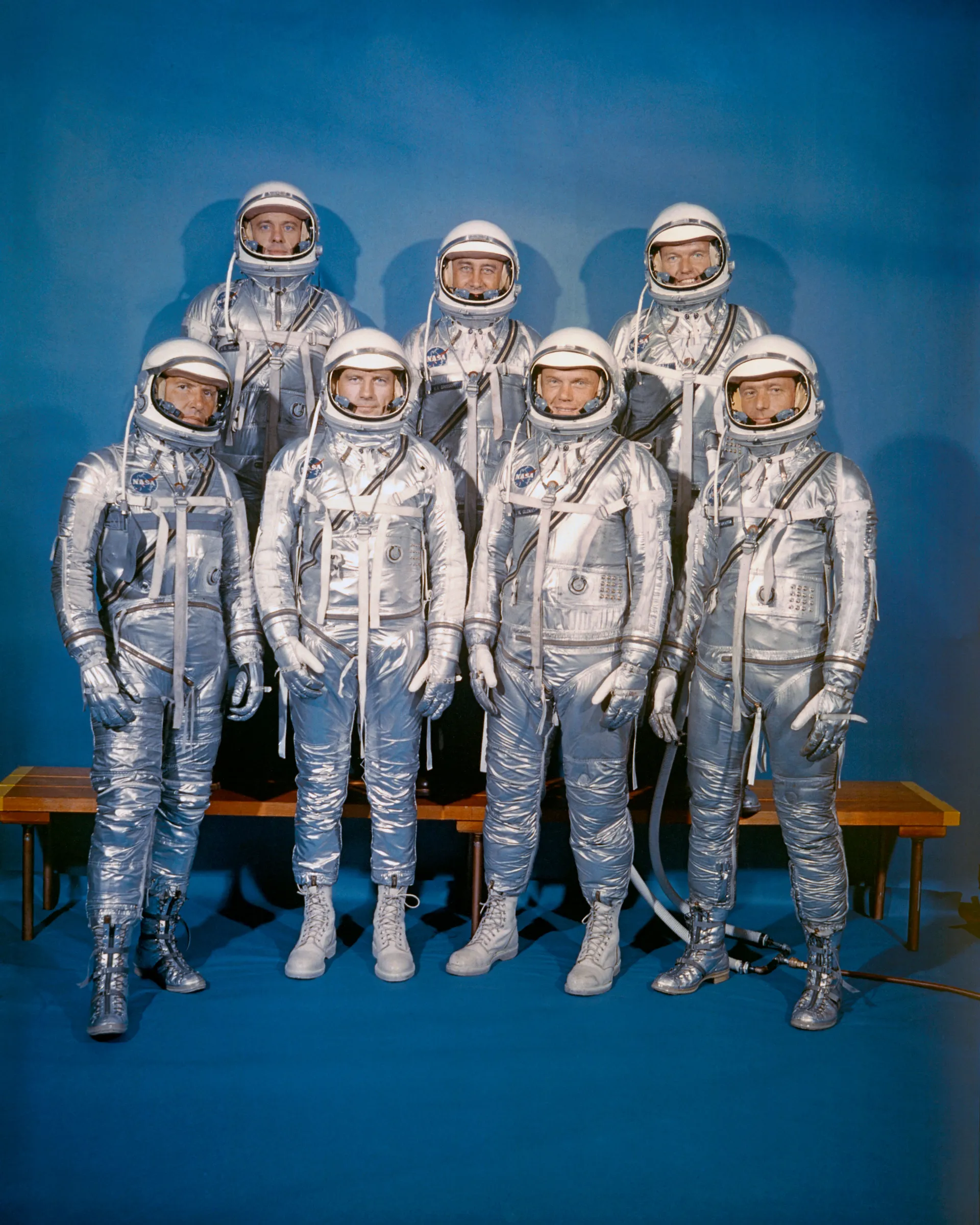 The seven Mercury astronauts in silver pressure suits, four seated in front and three standing behind.