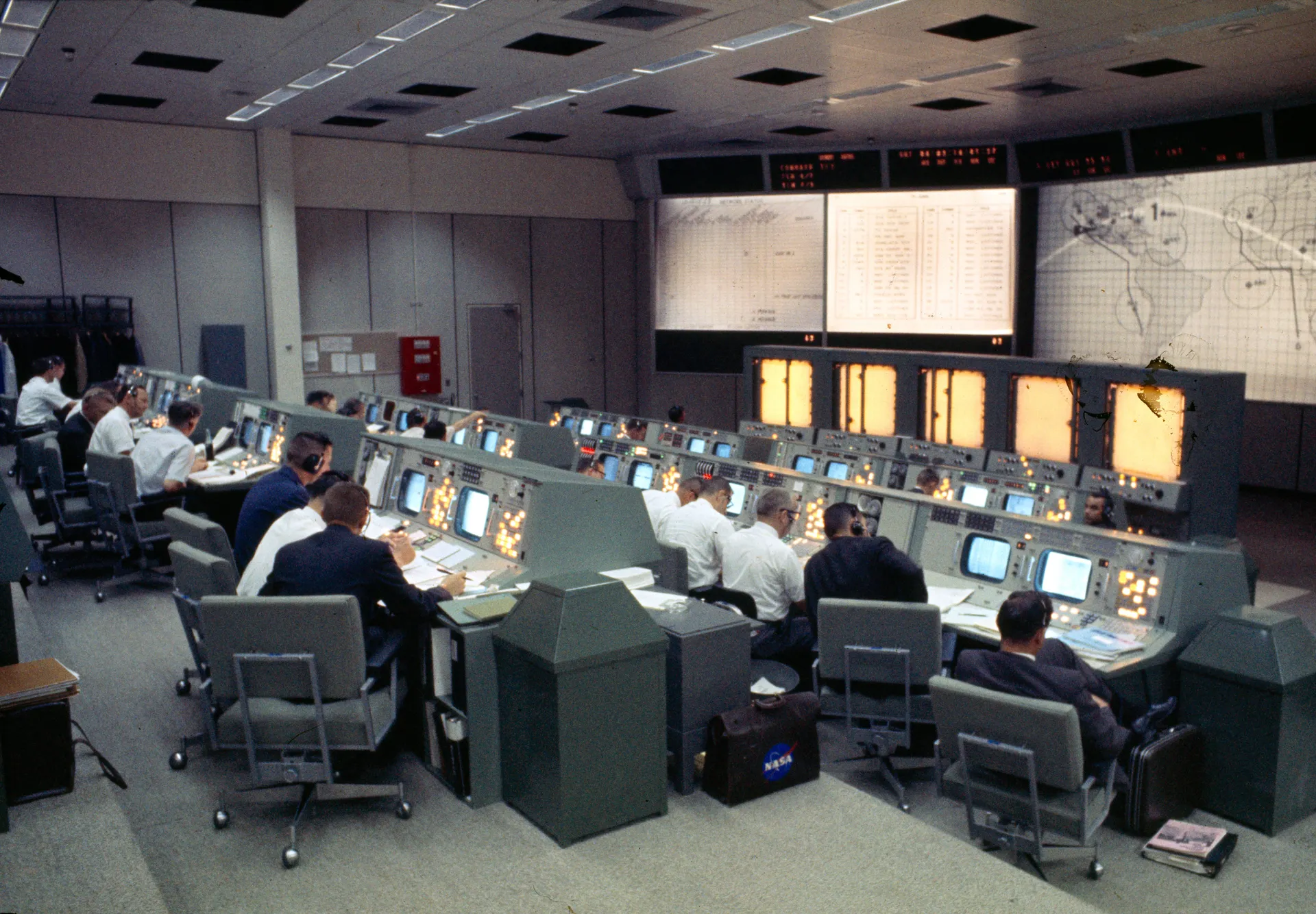 Rows of flight controllers at consoles in Houston’s Mission Control Center, with plot boards and a world map on the front wall.