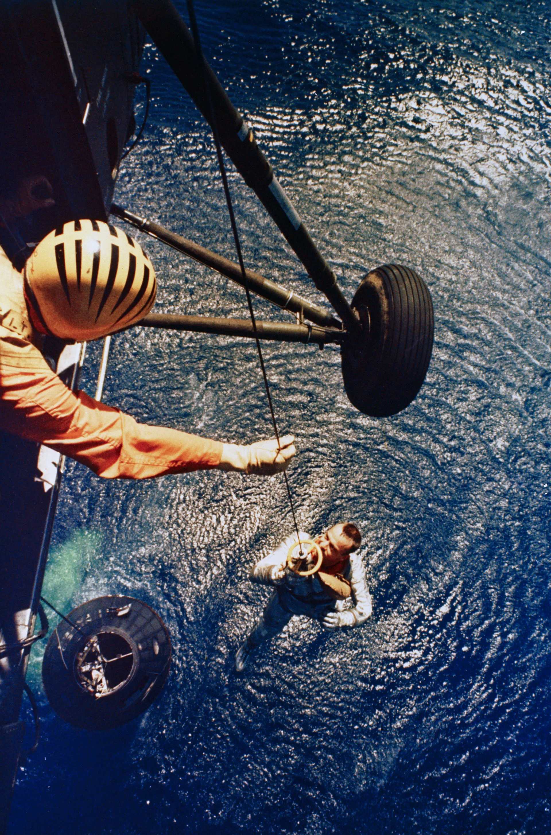 Seen from a helicopter, Alan Shepard rides a sling up from the ocean while Freedom 7 floats below.