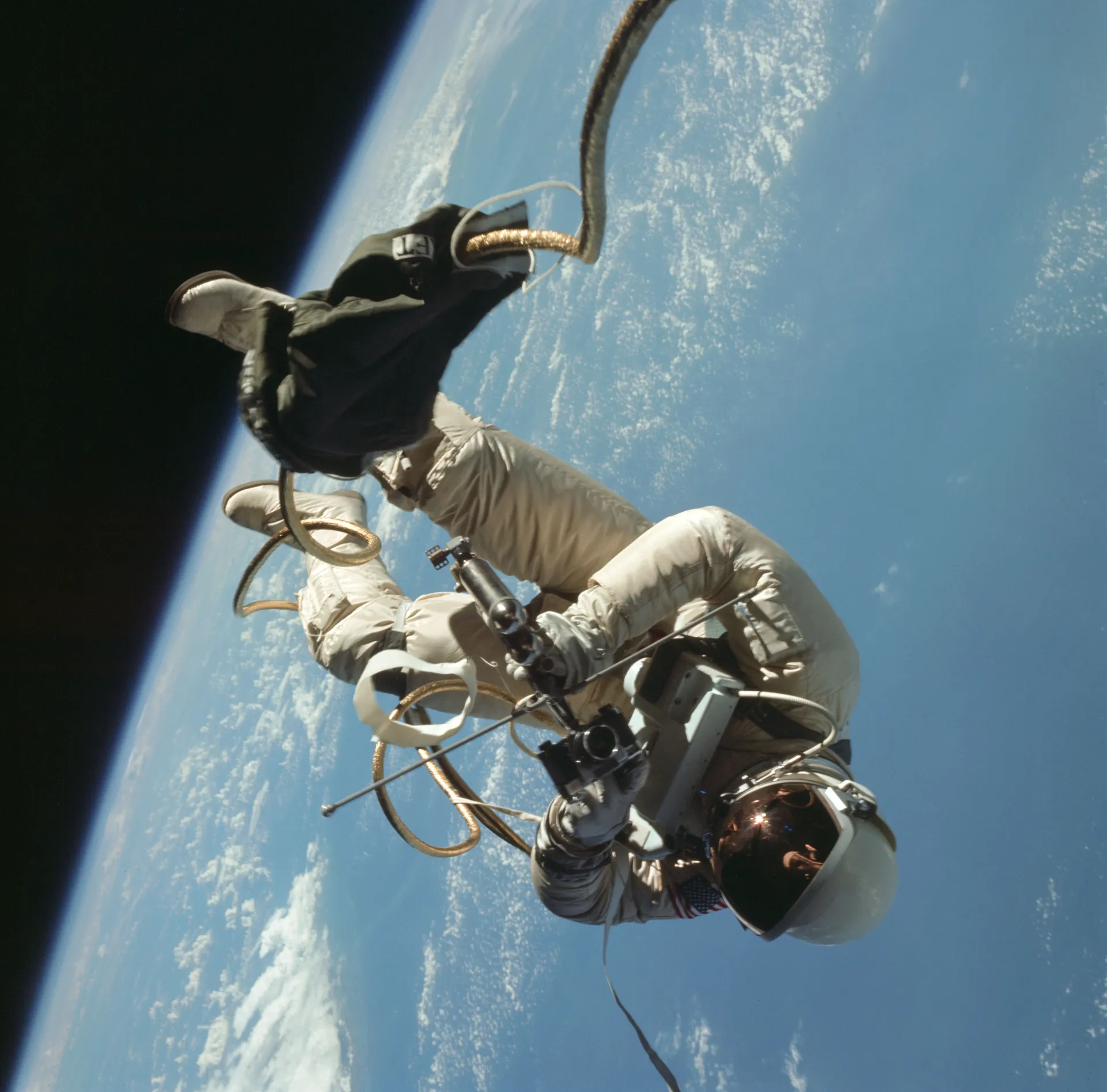 Ed White floats outside Gemini 4 above Earth, holding a hand-held maneuvering unit and attached by a gold umbilical.